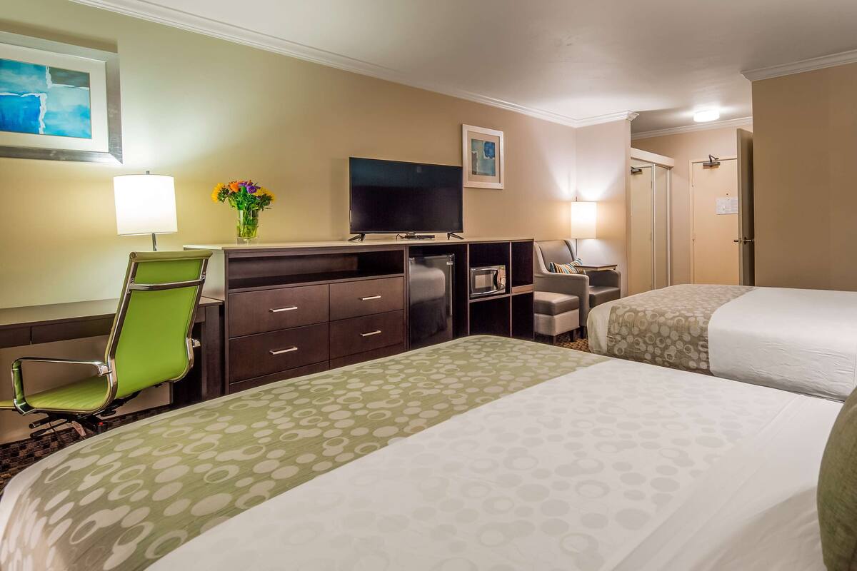 A hotel room with two beds, a desk with a flat-screen TV, a microwave, a green chair, and a lamp.