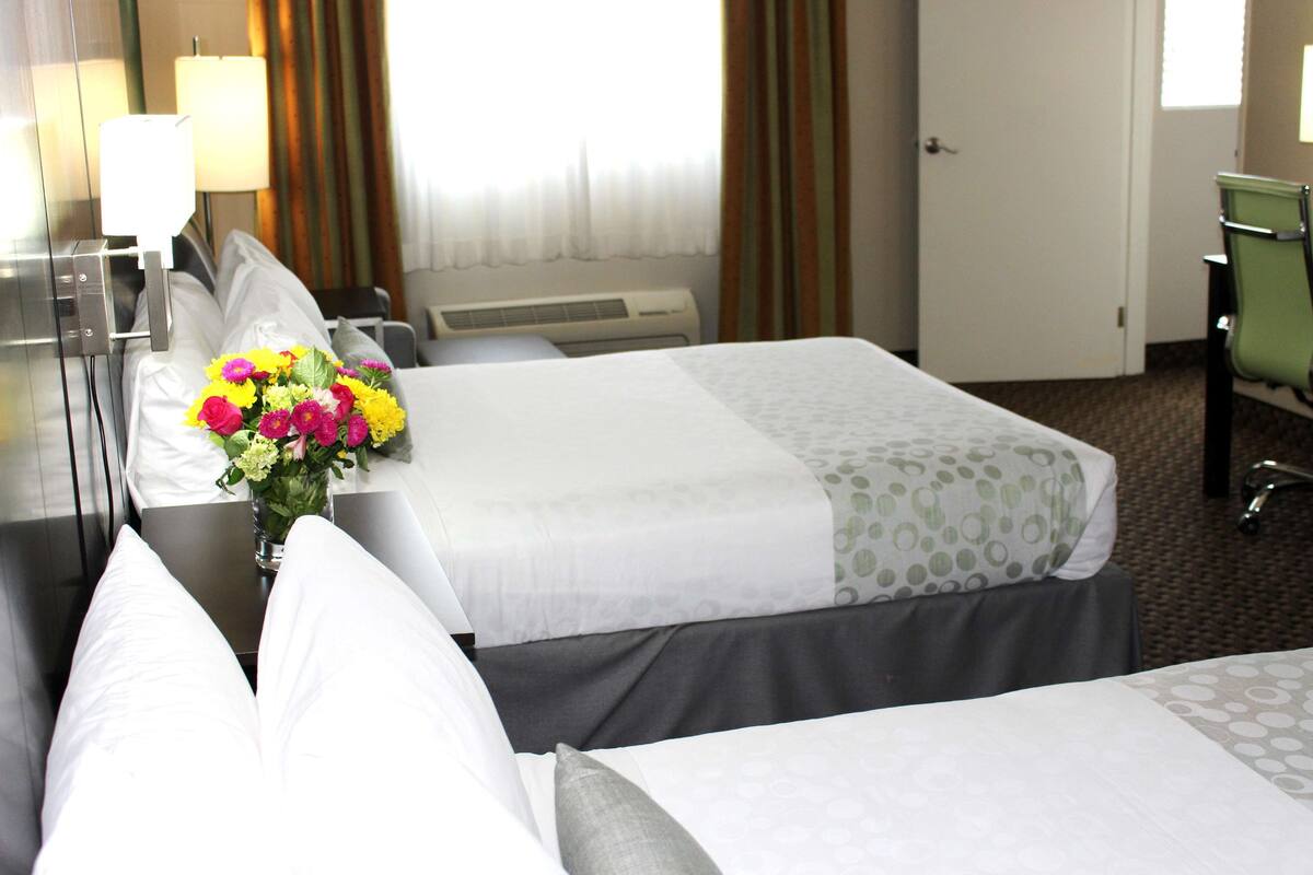 A hotel room with two beds, a desk with a lamp, a chair, and a window with curtains.