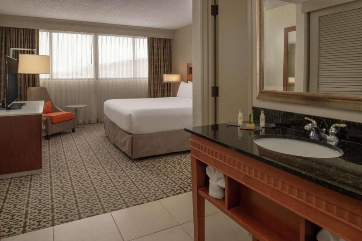 Una habitación de hotel con una cama, un escritorio con lámpara, una mesa redonda y un baño con lavabo y espejo.