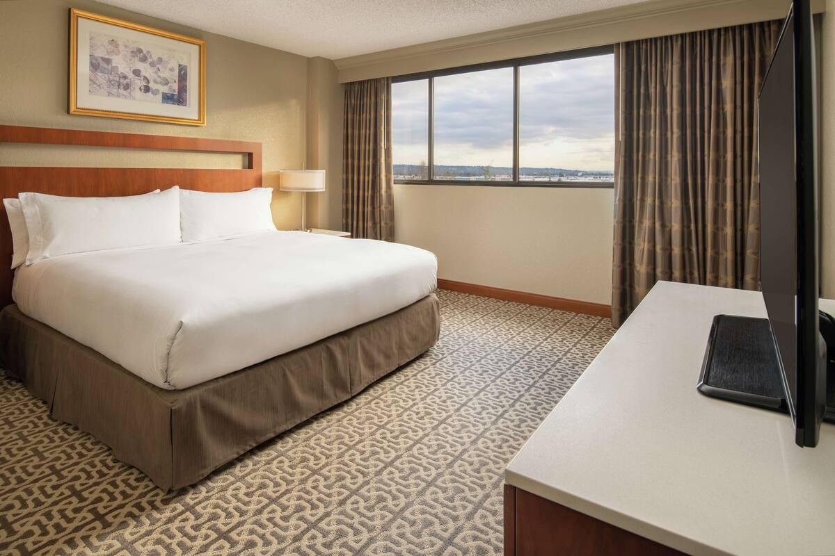 Habitación de hotel con una cama grande, un escritorio con computadora, una ventana con cortinas y un cuadro en la pared.