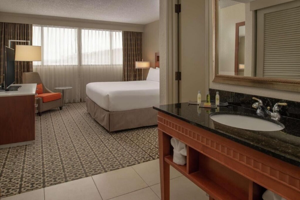 A hotel room with a bed, a desk with a lamp, a round table, a bathroom with a sink and mirror.