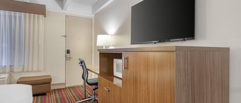 Habitación de hotel con cama, escritorio con silla, televisión, lámpara y armario.