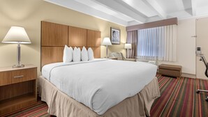 A hotel room with a large bed, wooden headboard, two bedside lamps, a small table, a chair, a window with curtains, and a bathroom with a shower and toilet. - Best Western El Rancho Palacio (Roswell)