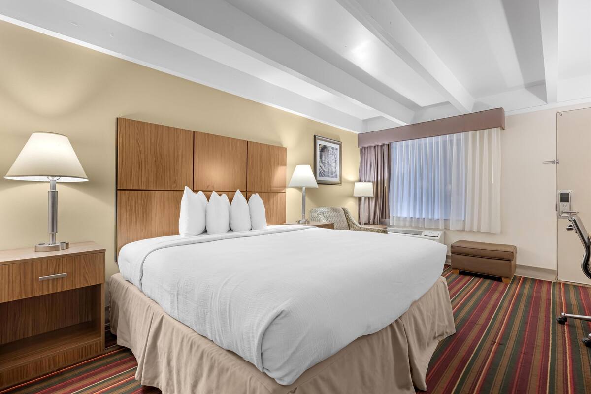 A hotel room with a large bed, wooden headboard, two bedside lamps, a small table, a chair, a window with curtains, and a bathroom with a shower and toilet.
