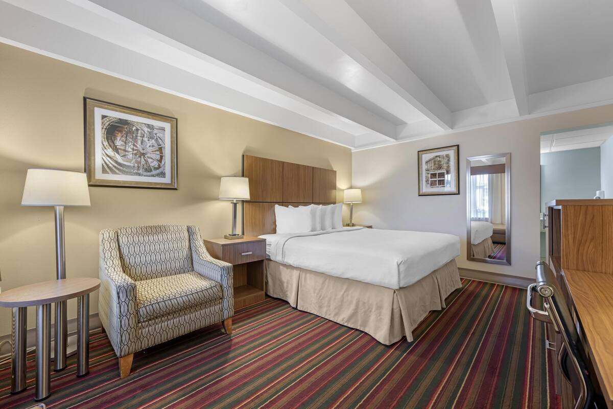 A hotel room with a bed, a patterned armchair, a small round table, and a striped carpet.
