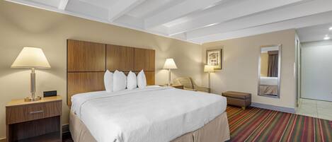 A hotel room with a large bed, two bedside tables with lamps, a wooden headboard, a striped carpet, and a small seating area with a chair and table.