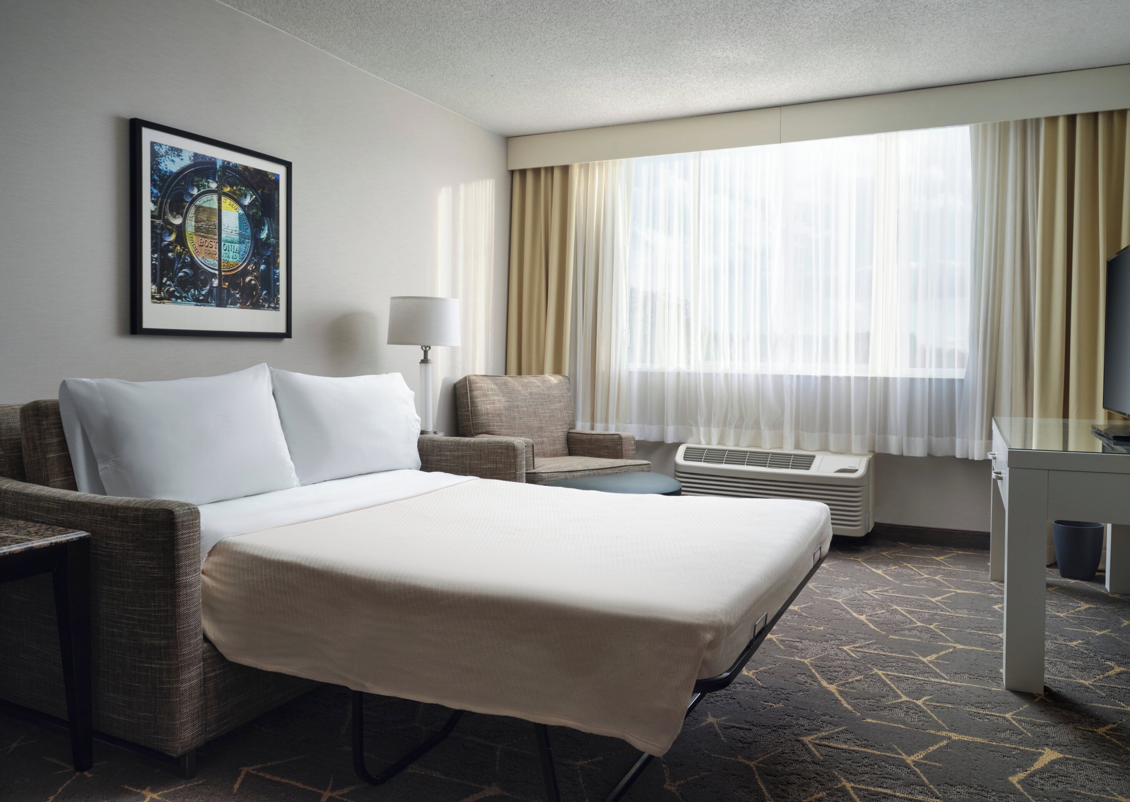 A hotel room with a bed, a desk, a chair, a lamp, a television, and a window with curtains.
