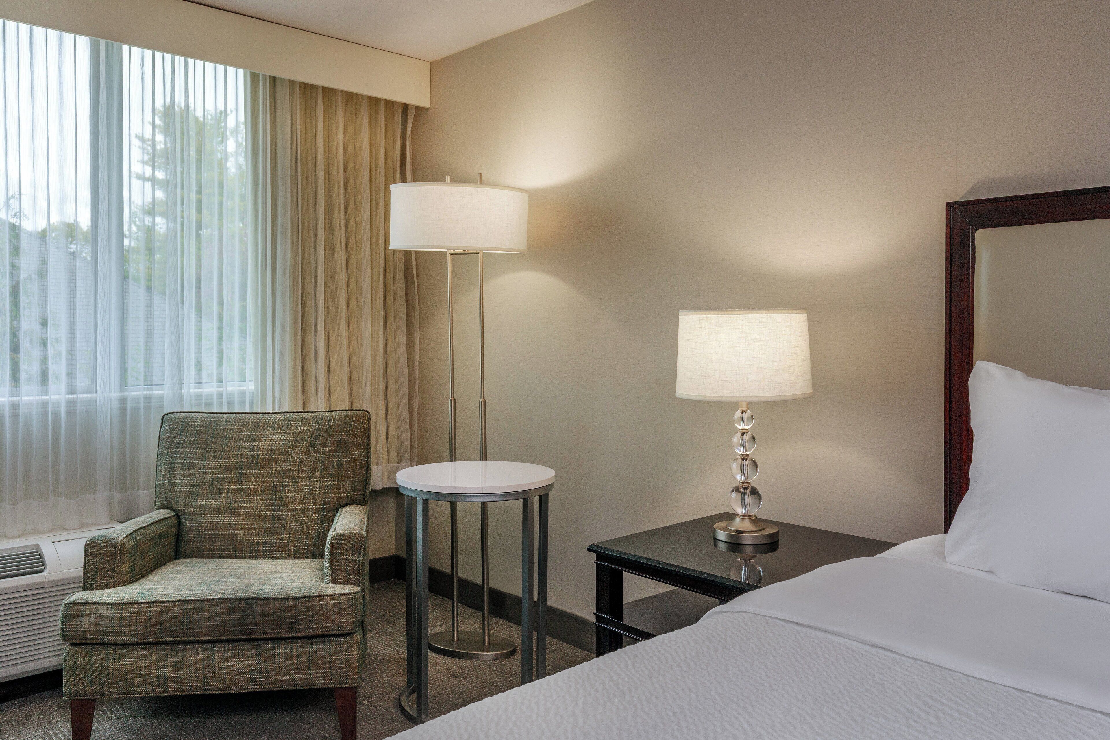 A hotel room with a bed, a chair, a small table, and a lamp.