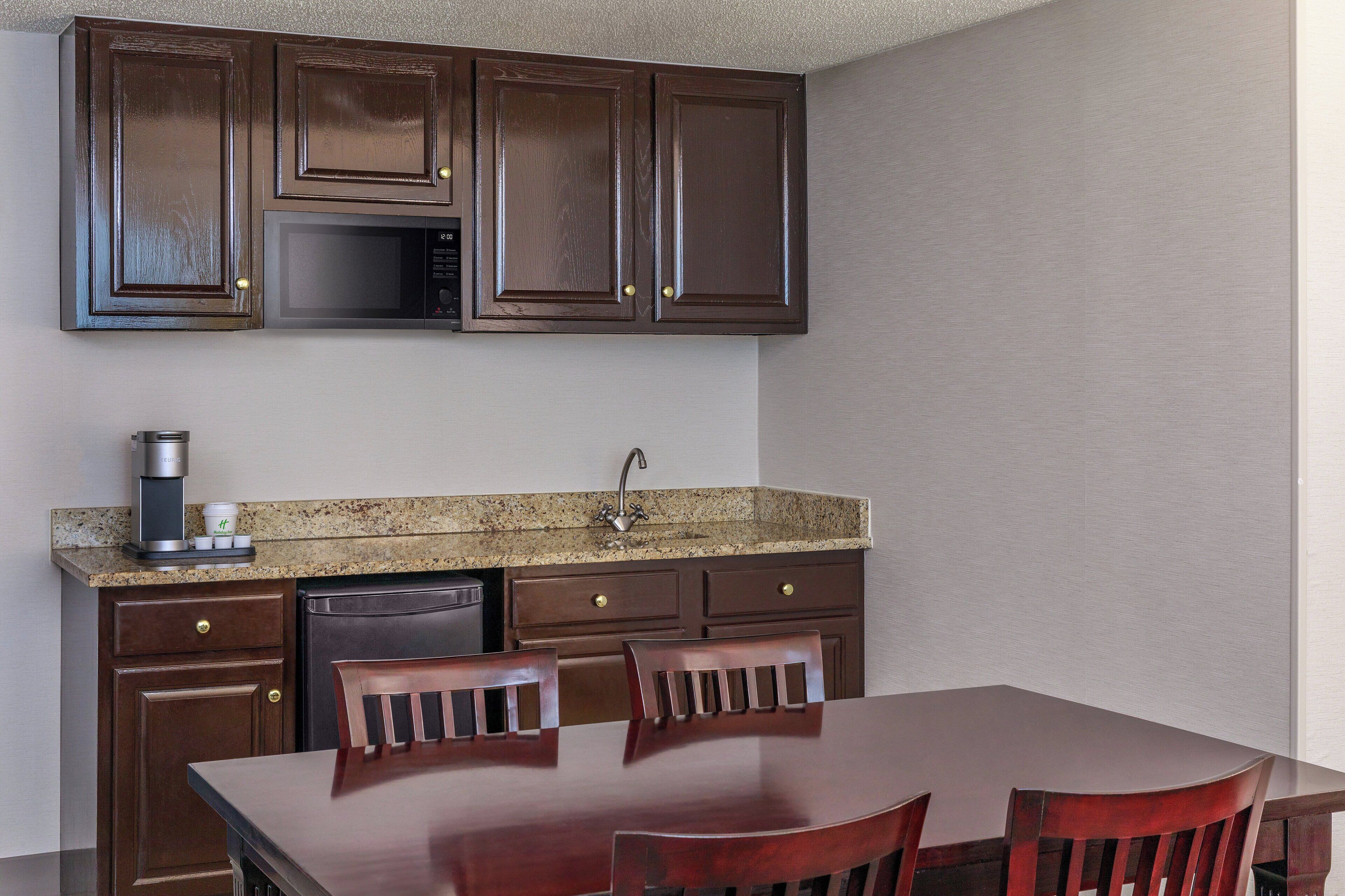 A compact kitchen with dark wooden cabinets, a granite countertop, a built-in microwave, a coffee maker, and a dishwasher. There is a wooden dining table with two chairs.