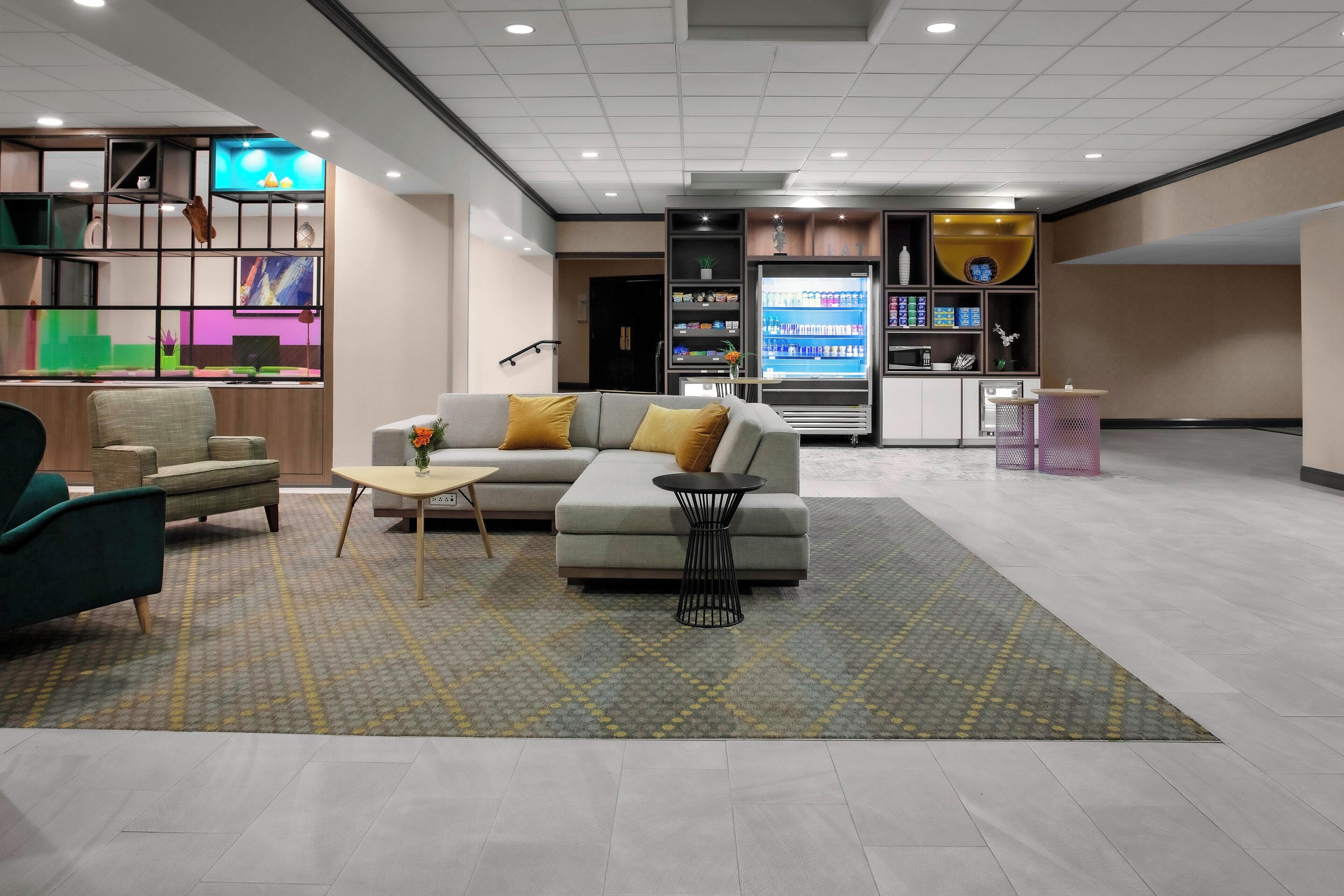 Eine moderne Lobby mit einer Couch, Sesseln, einem Couchtisch, einem mit Getränken gefüllten Kühlschrank und einem dekorativen Regal mit Pflanzen.