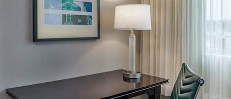 Habitación de hotel con una mesa de madera oscura, una lámpara, un cuadro en la pared y una silla tapizada en verde.