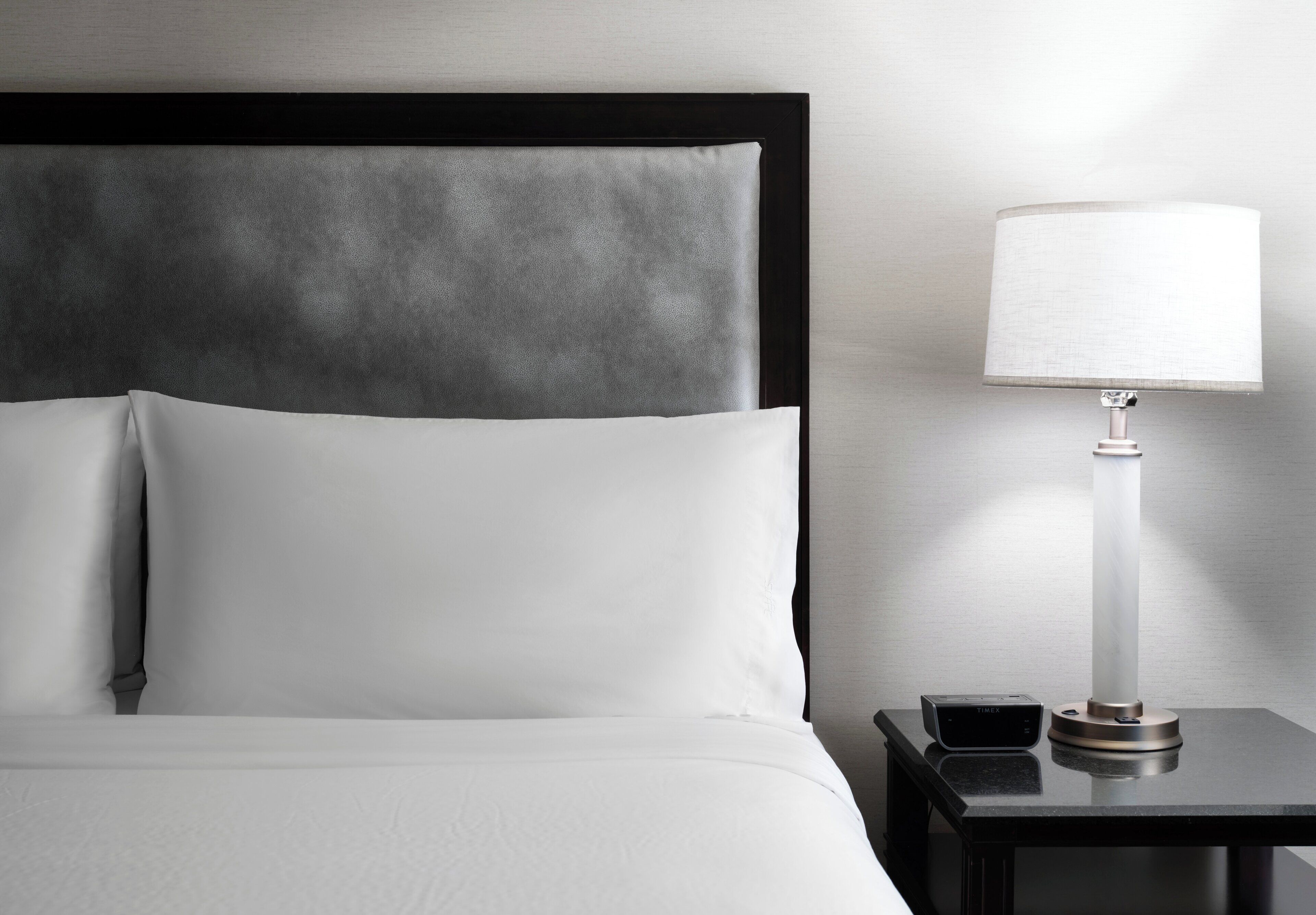 A neatly made bed with white linens, a dark headboard, and a bedside table with a lamp and a small electronic device.