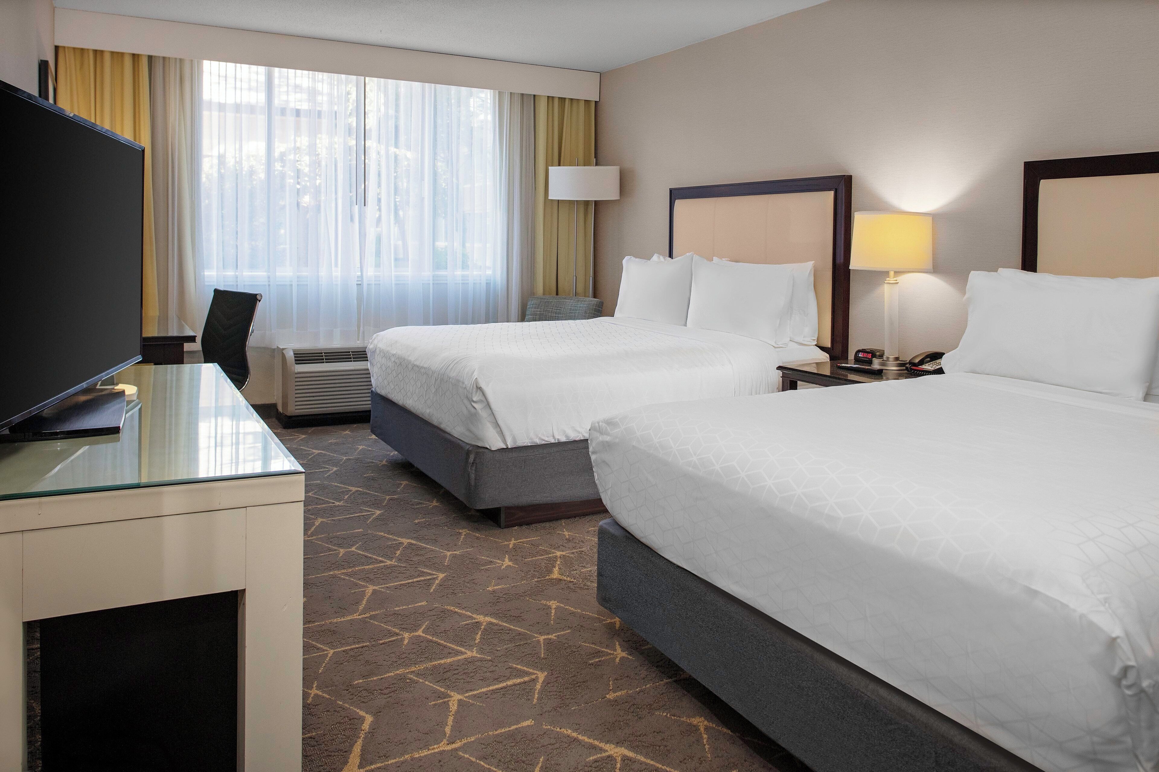 A hotel room with two beds, a desk with a TV, a lamp, and a clock on the nightstand.
