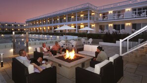 A poolside area with outdoor seating, a fire pit, and a building with multiple balconies and outdoor lighting. - Ocean House Hotel at Bass Rocks (Gloucester)