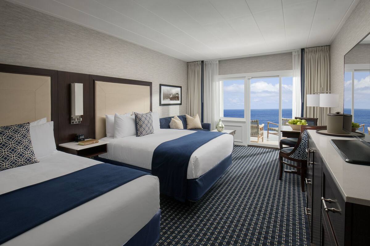 Habitación de hotel con dos camas, un escritorio con lámpara, una silla y vista al océano.