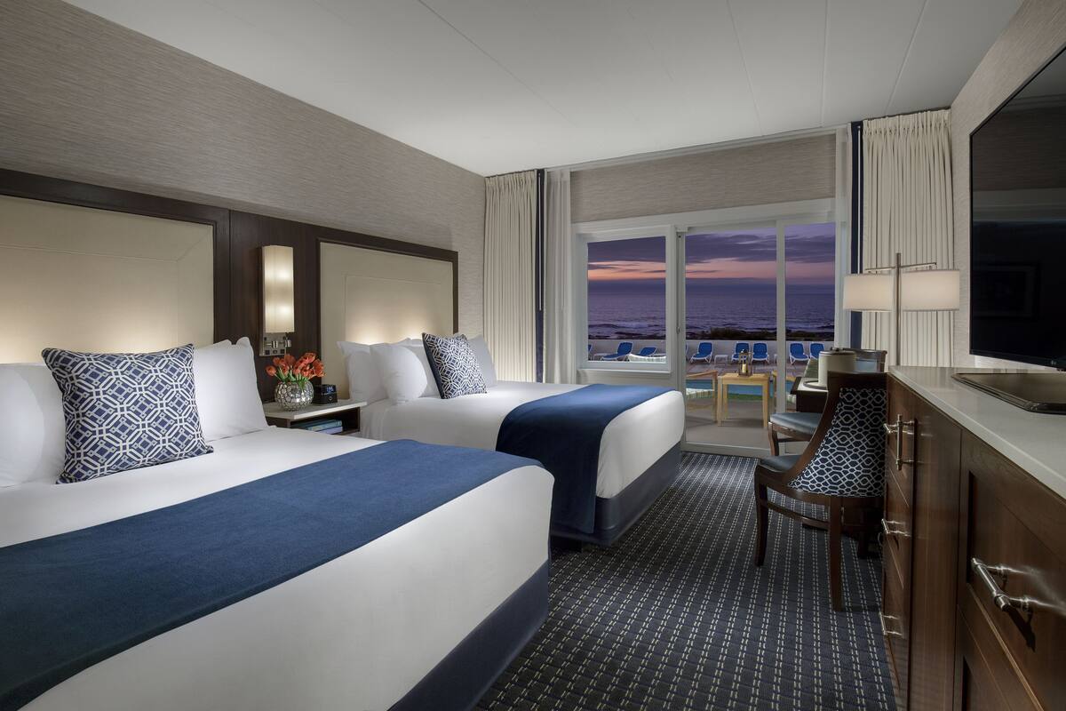 Habitación de hotel con dos camas, un escritorio, una silla y vista al océano al atardecer.