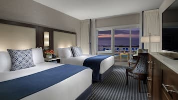 Habitación de hotel con dos camas, un escritorio, una silla y vista al océano al atardecer.