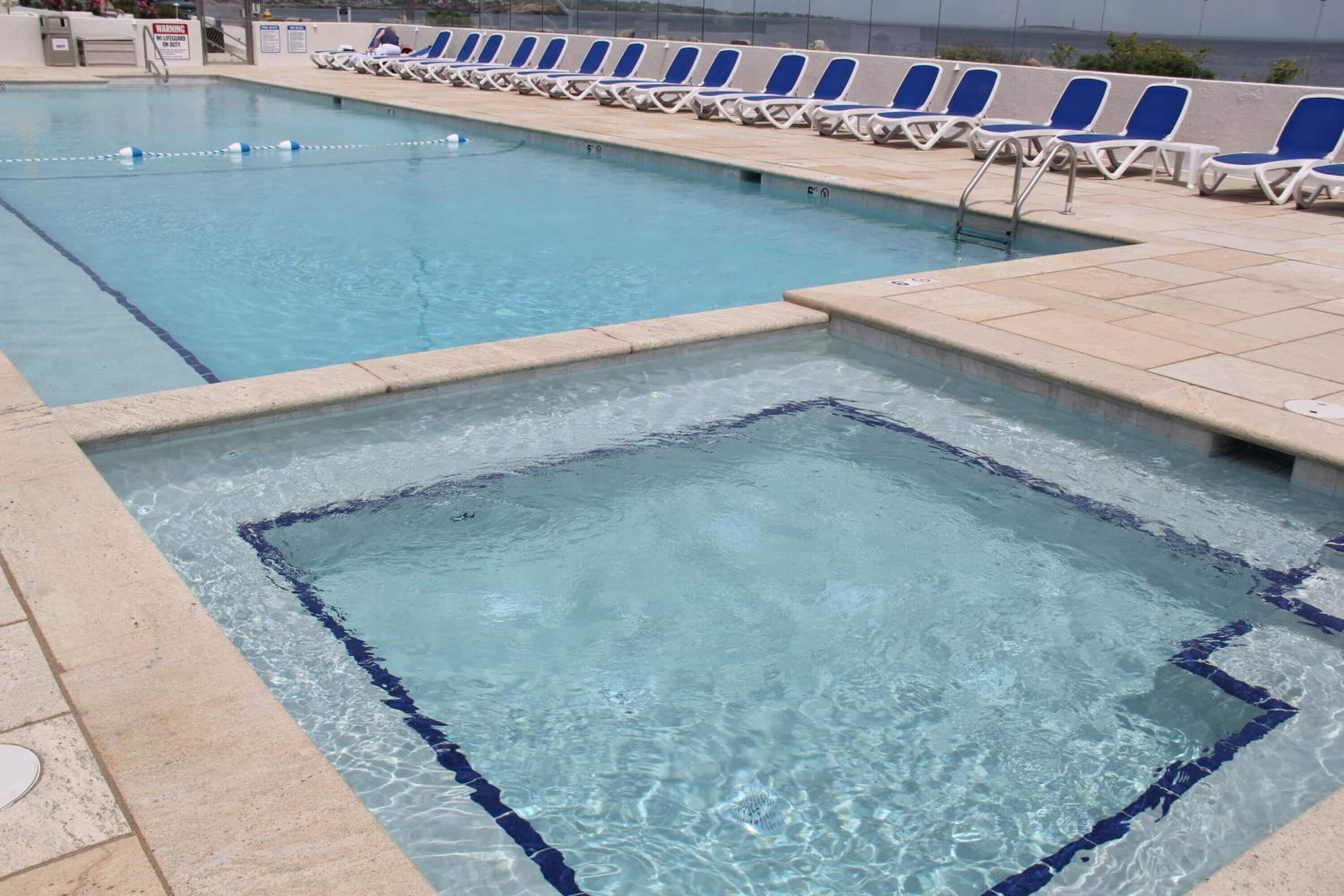 A swimming pool with clear blue water, a diving board, and a row of blue and white lounge chairs along the edge.