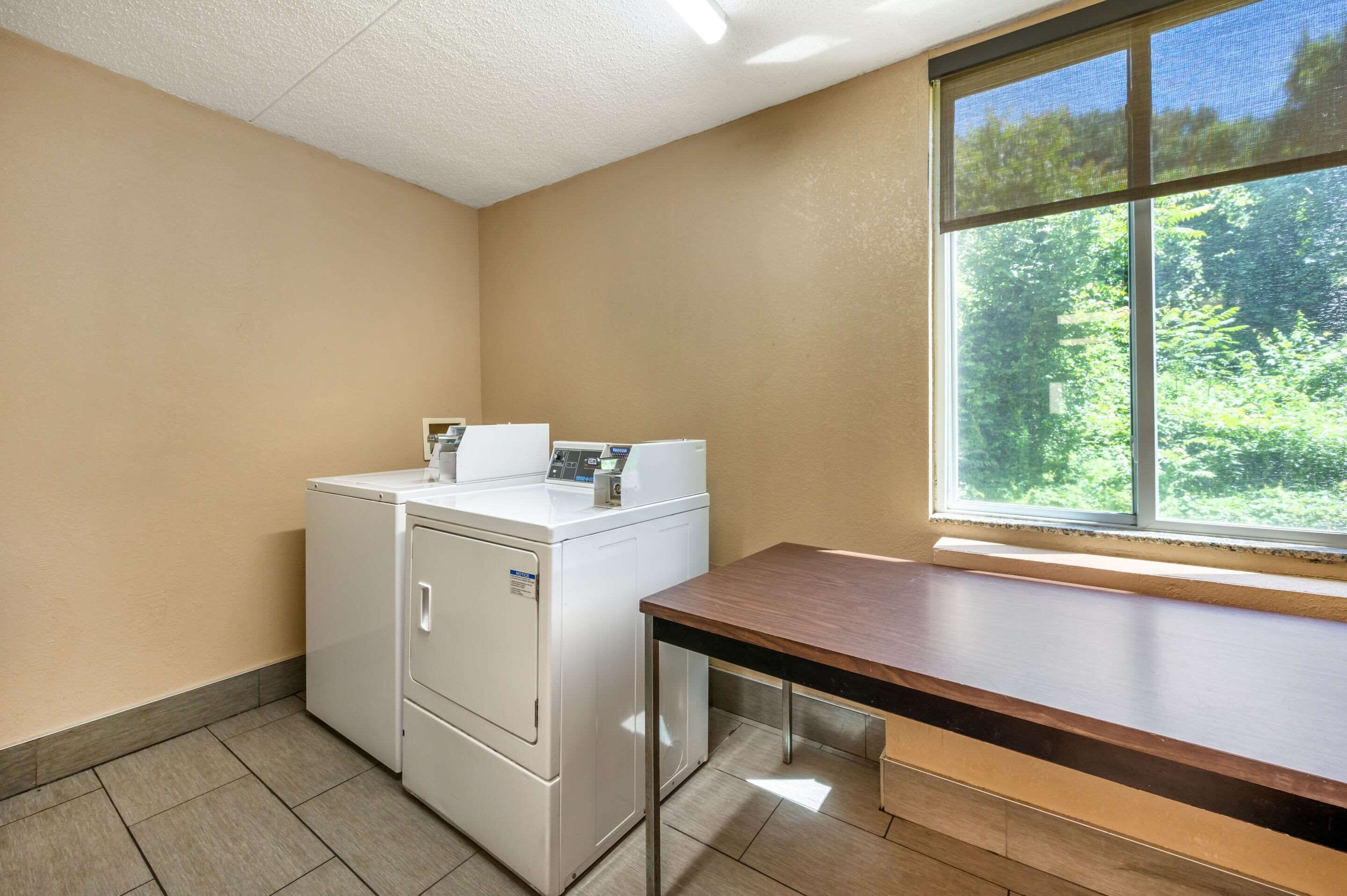 A laundry room with a washing machine and dryer, a wooden bench, and a window with a view of greenery.