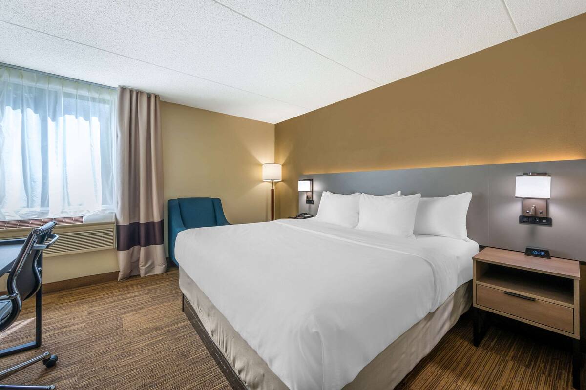 Hotellihuone, jossa on suuri sänky, työpöytä, tuoli, lamppu ja yöpöytä, jossa on kello.