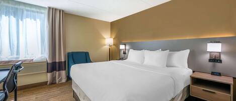 A hotel room with a large bed, a desk, a chair, a lamp, and a nightstand with a clock.