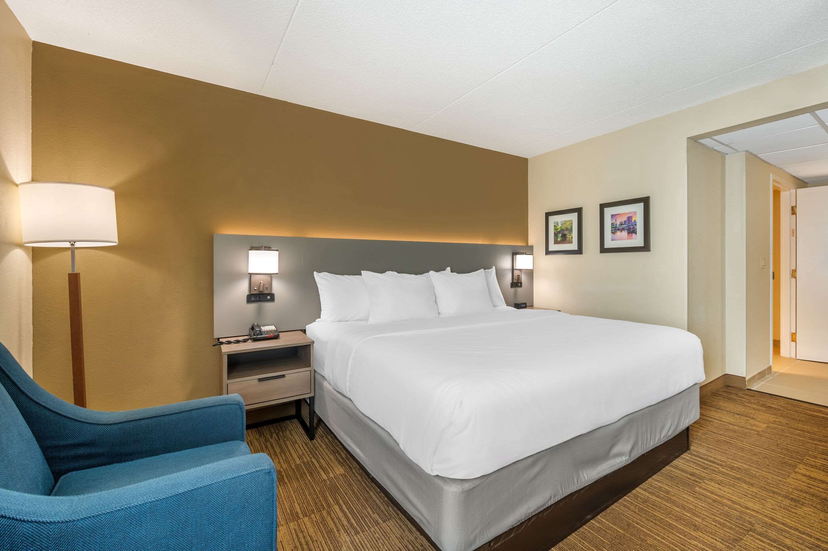 A hotel room with a large bed, a nightstand with a lamp, a blue armchair, and two framed pictures on the wall.