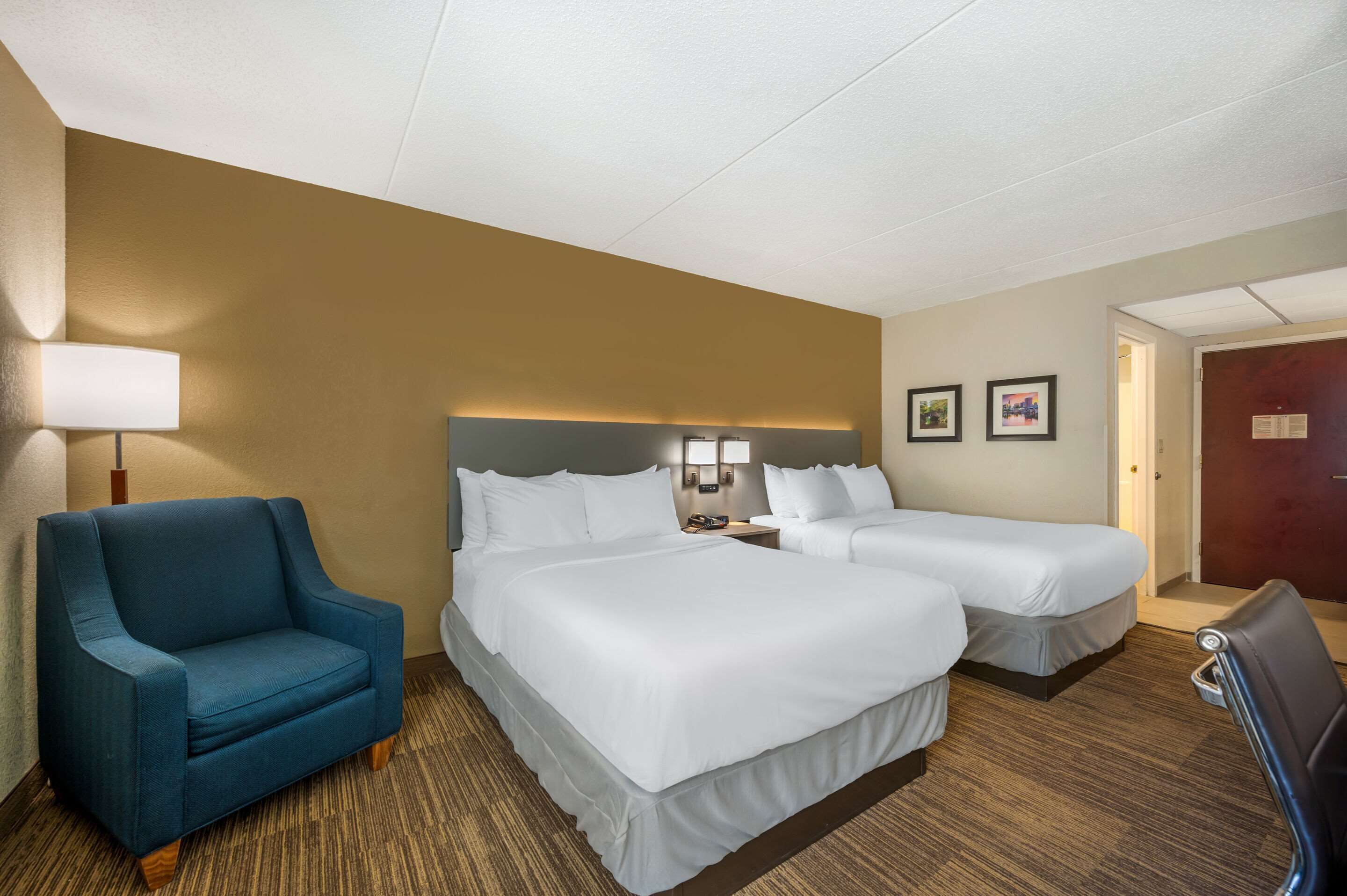 A hotel room with two beds, a blue armchair, a lamp, and a door.