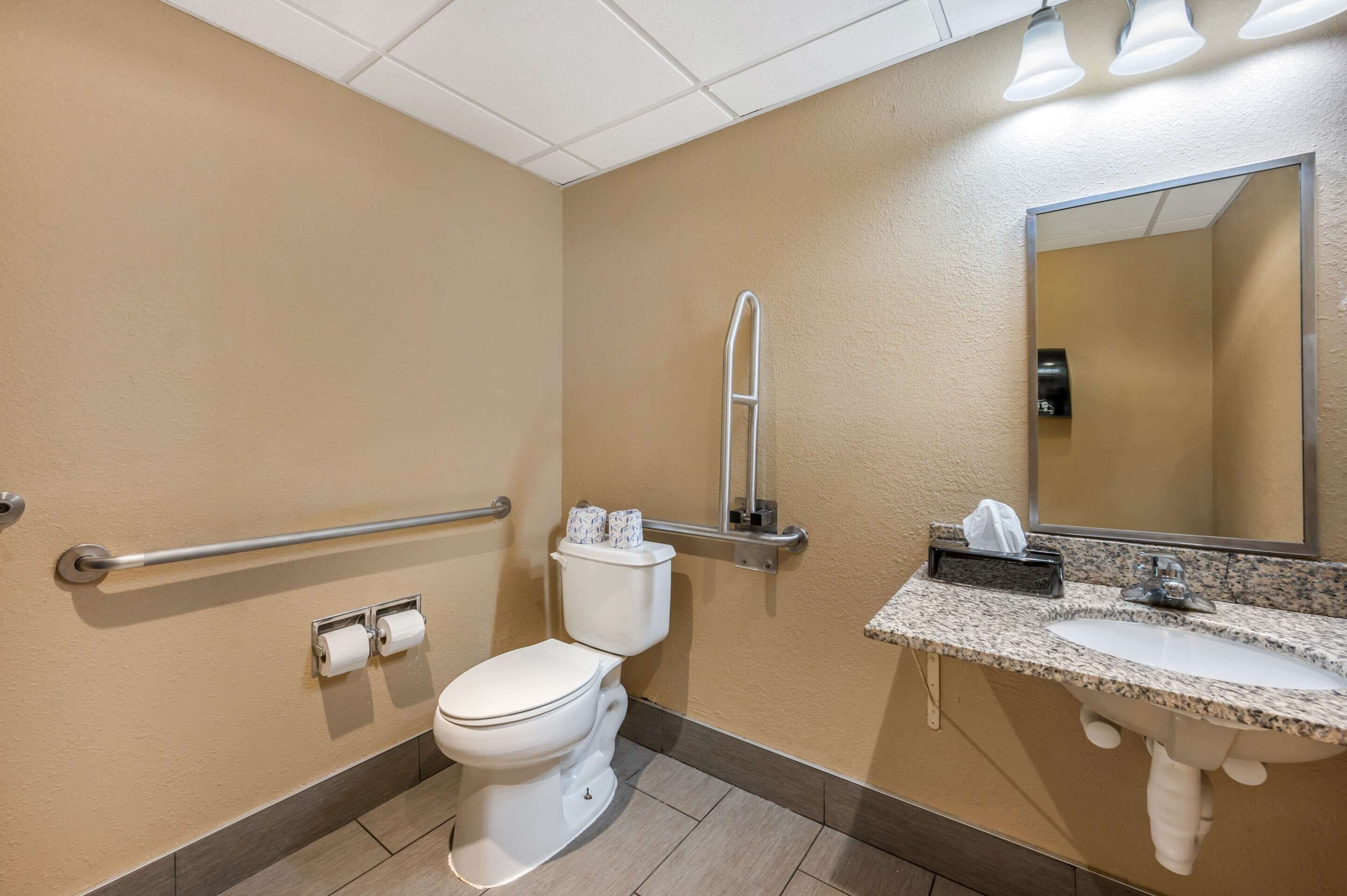 A bathroom with a toilet, handrail, towel dispenser, and sink.