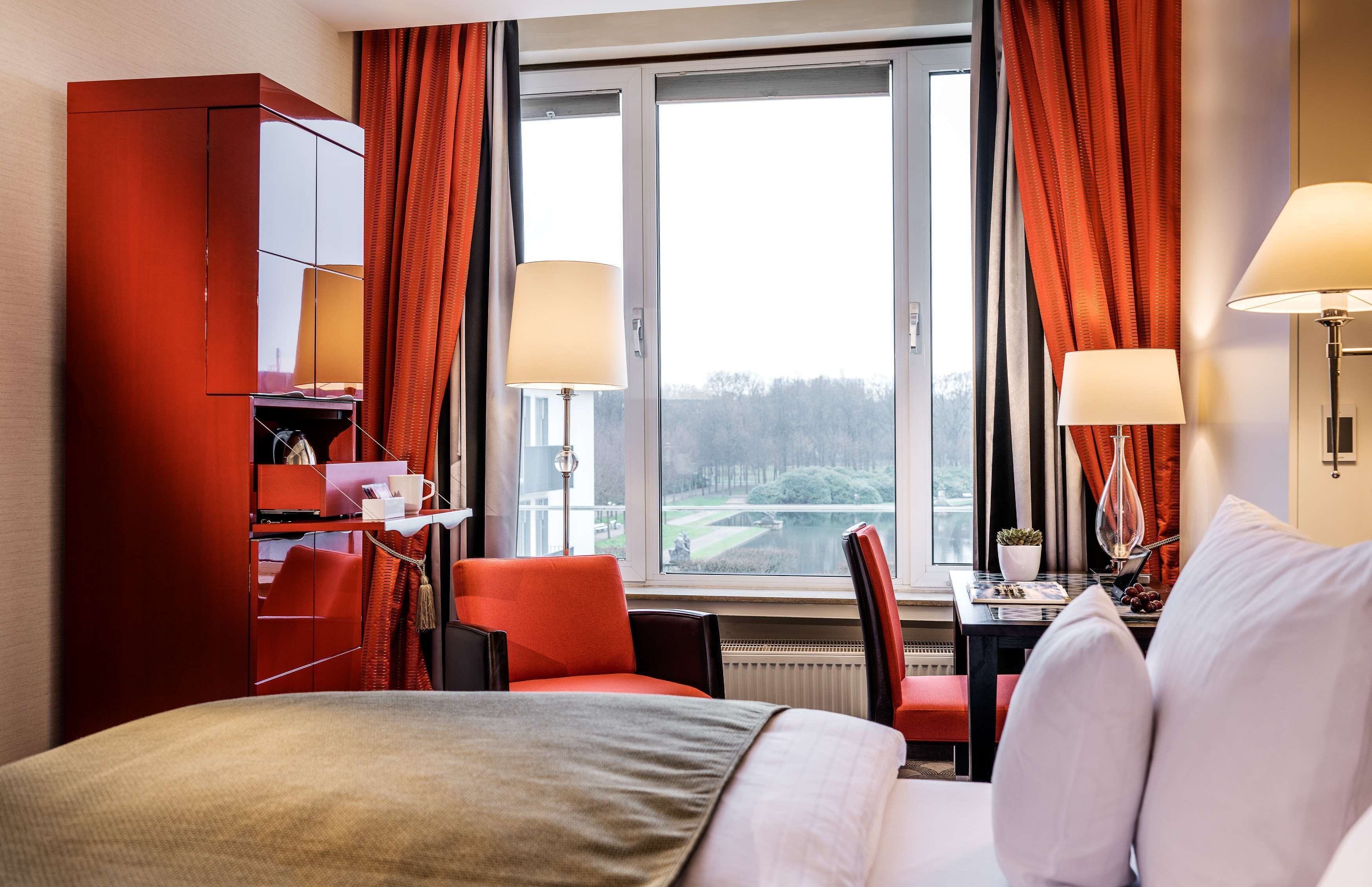 A hotel room with a bed, a desk, a chair, a red wardrobe, and a window with curtains.