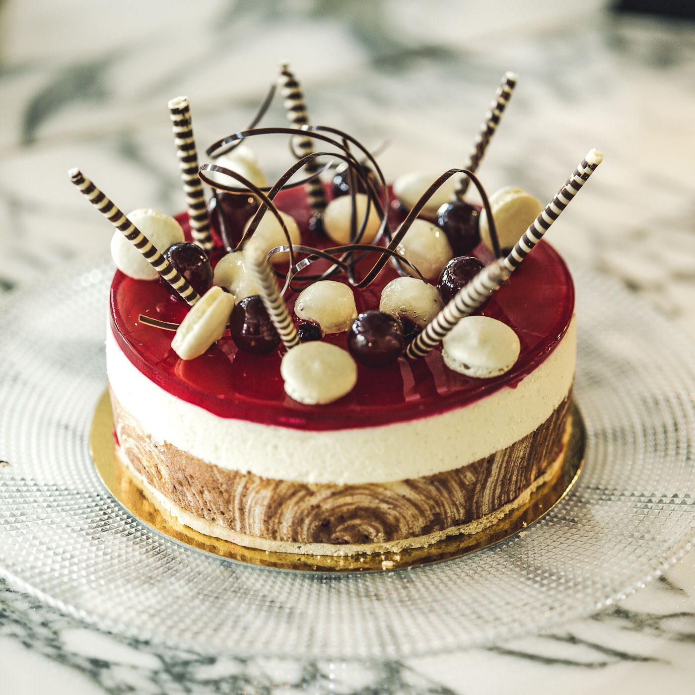 A layered cake with a red jelly topping, white cream layer, and a chocolate base, decorated with cherries and chocolate curls.