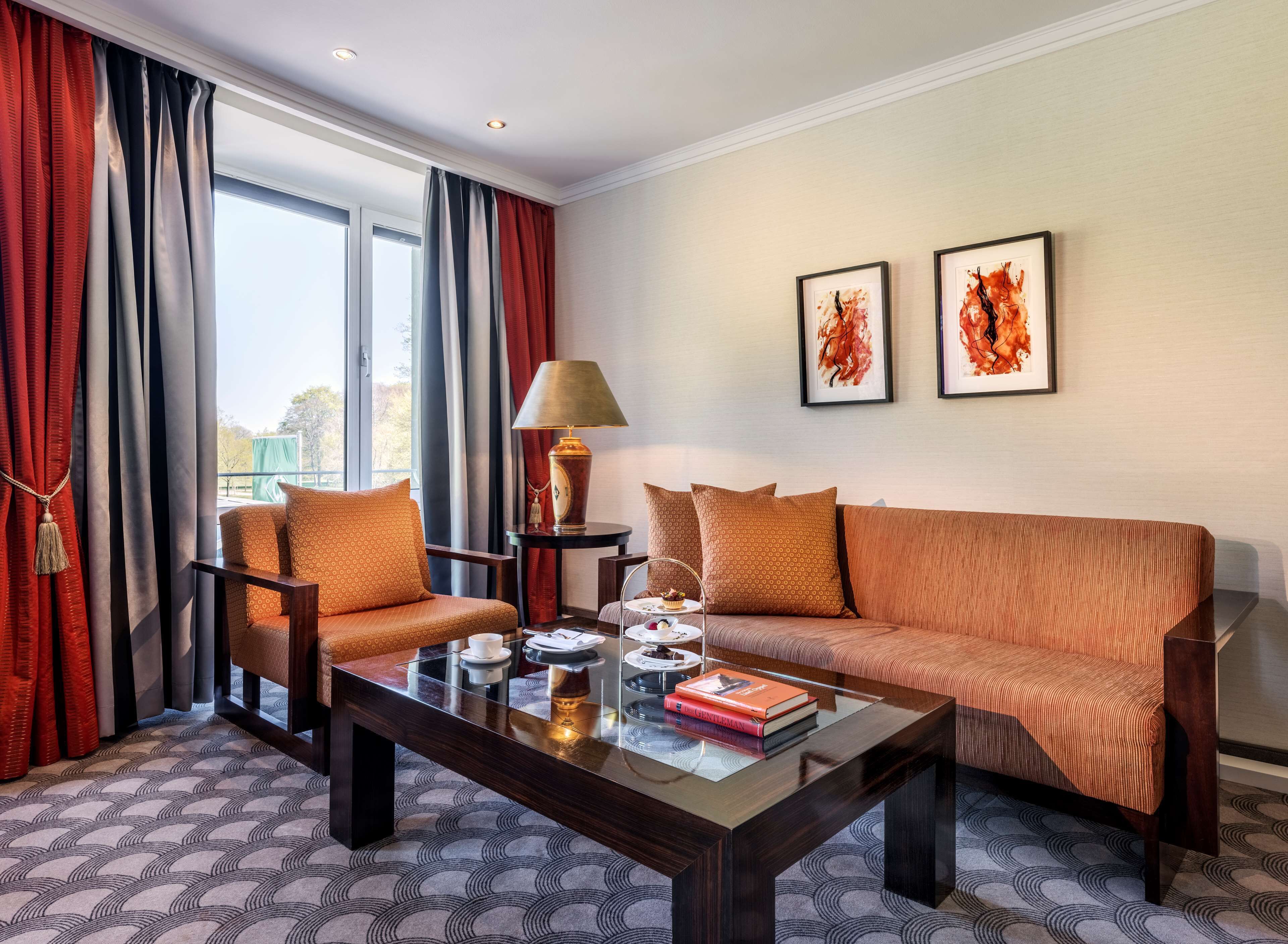 Ein Hotelzimmer mit Sofa, Sessel und Couchtisch. Eine Lampe, ein Tablett mit Teekanne und Tassen sowie zwei gerahmte Kunstwerke an der Wand sind zu sehen.