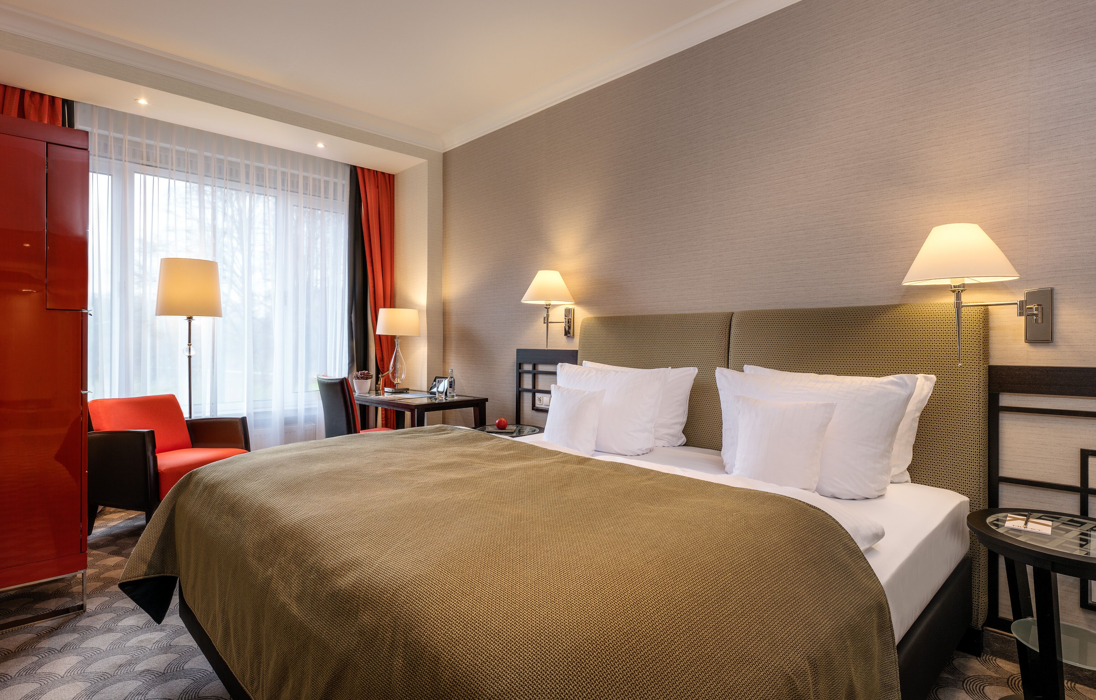 Ein Hotelzimmer mit einem großen Bett, zwei Nachttischlampen, einem roten Sessel, einem kleinen Tisch und einem fenster mit Vorhängen.