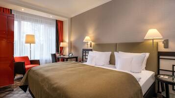 Ein Hotelzimmer mit einem großen Bett, zwei Nachttischlampen, einem roten Sessel, einem kleinen Tisch und einem fenster mit Vorhängen.