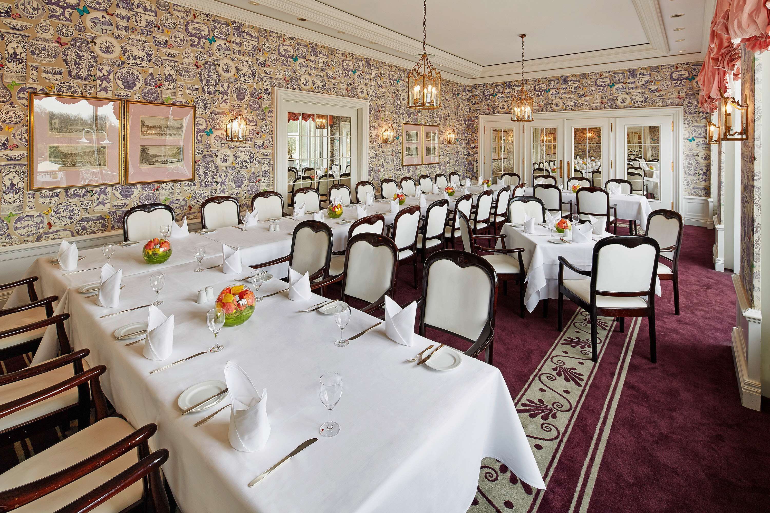 A formal dining room with multiple tables set for service, white tablecloths, and a patterned wallpaper design.