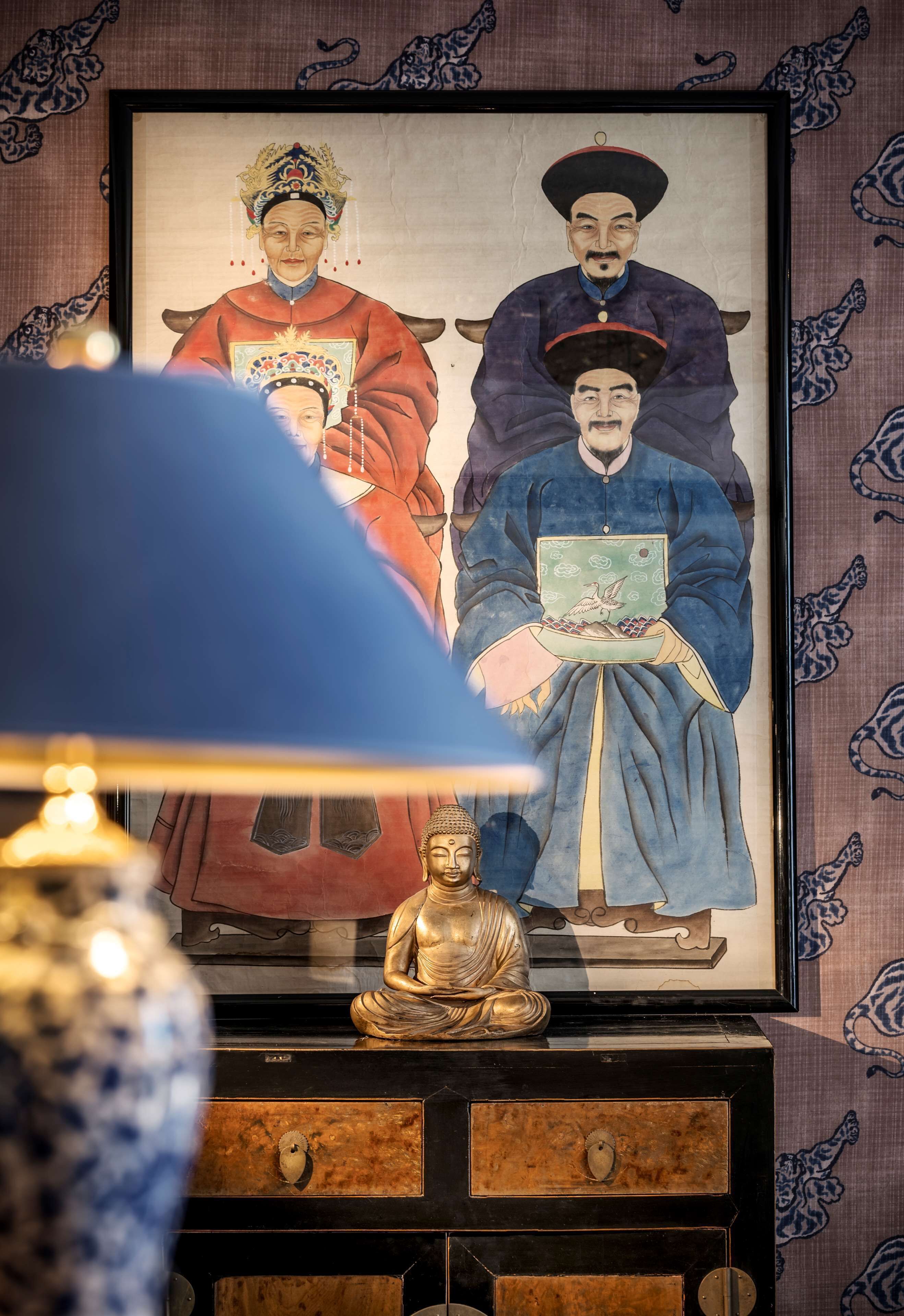 Ein gerahmtes Gemälde von historischen Persönlichkeiten an einer Wand, eine Buddha-Statue auf einem Holztisch und eine blaue Lampe im Vordergrund.