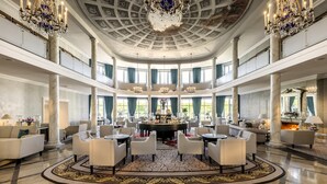 Un amplio vestíbulo con una mesa redonda central, varios conjuntos de asientos, candelabros y grandes ventanales que ofrecen vistas al exterior.