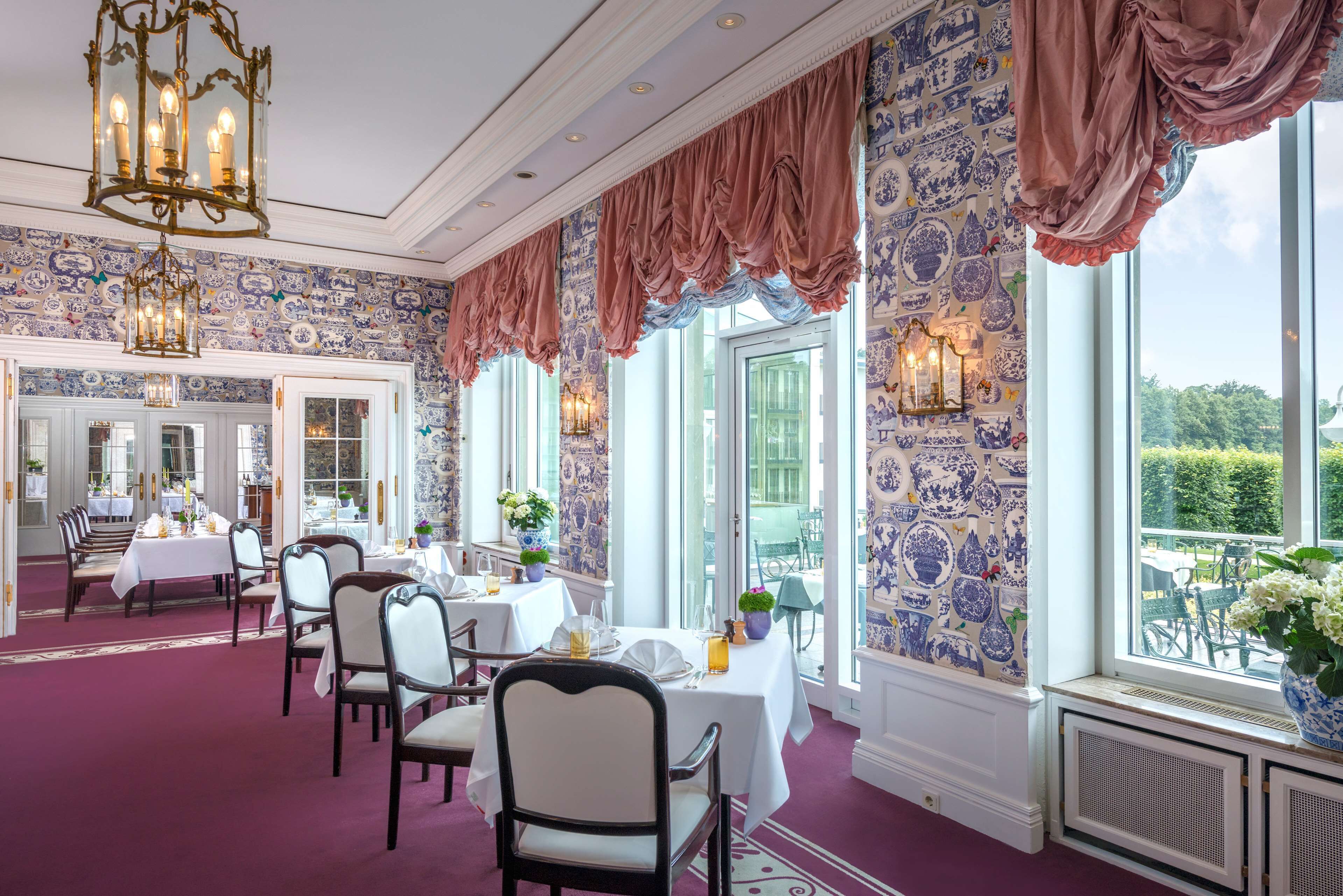 A dining area with tables set for a meal, a chandelier, and a view of the outdoors through large windows.