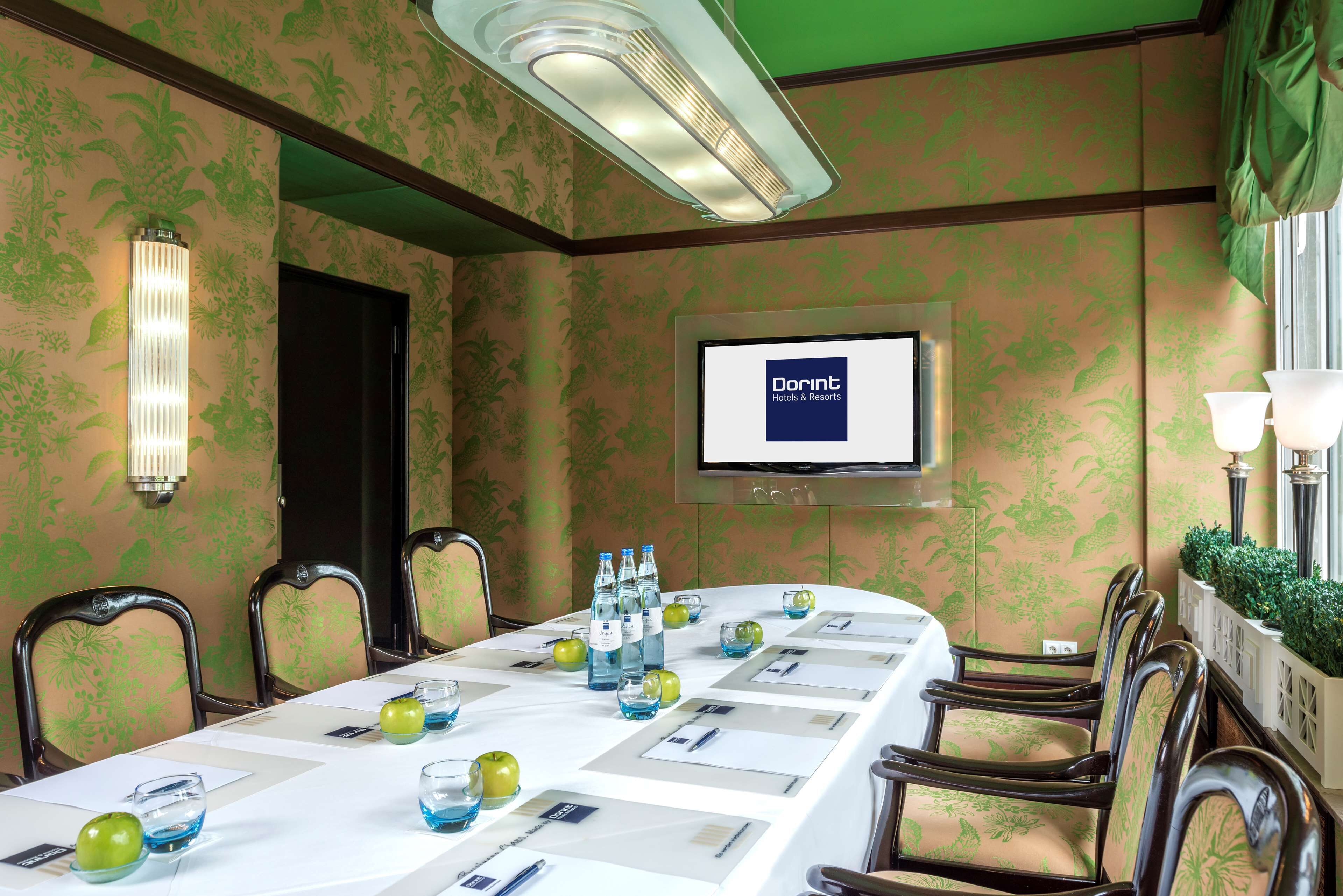 A conference room with a long table, chairs, a projector screen, and a wall-mounted TV displaying 'Dorint Hotels & Resorts'.