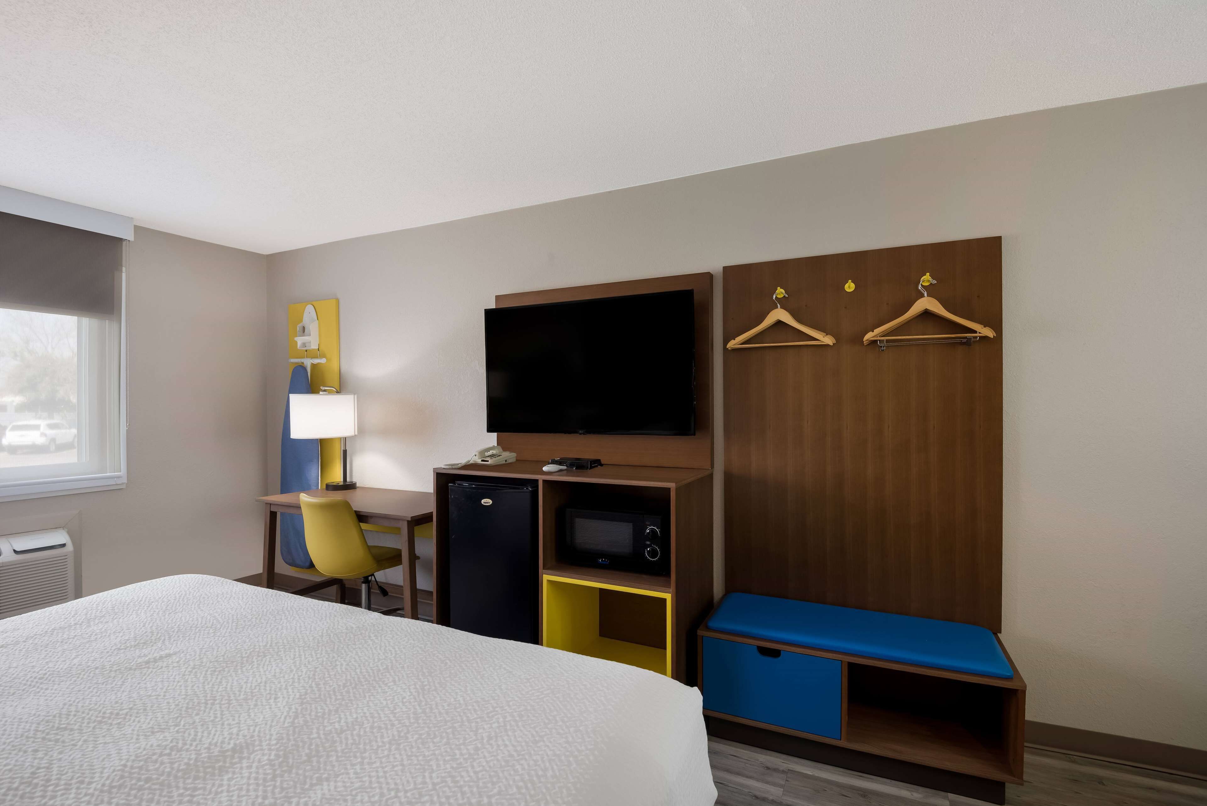 A hotel room with a bed, a desk with a computer, a television, a mini-fridge, a microwave, and a blue bench.