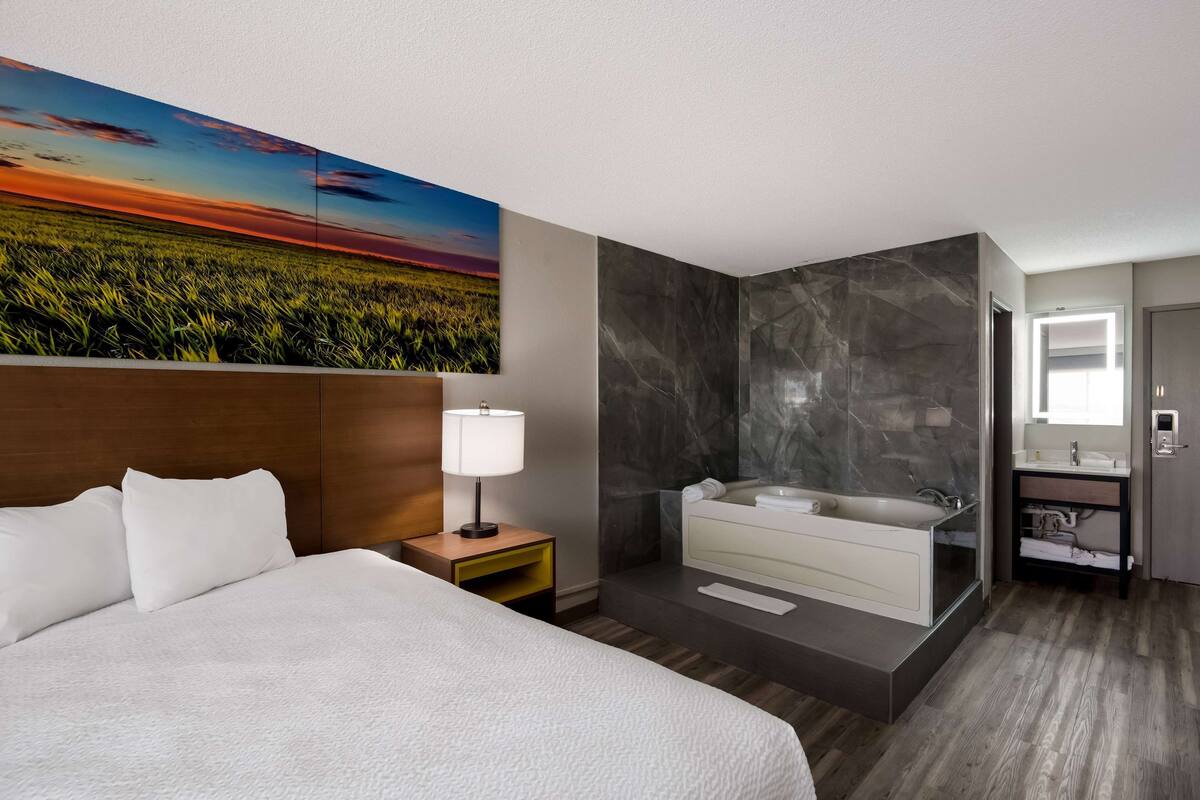A modern hotel room with a large bed, a nightstand, a wall-mounted painting, a marble bathroom, and a wooden floor.