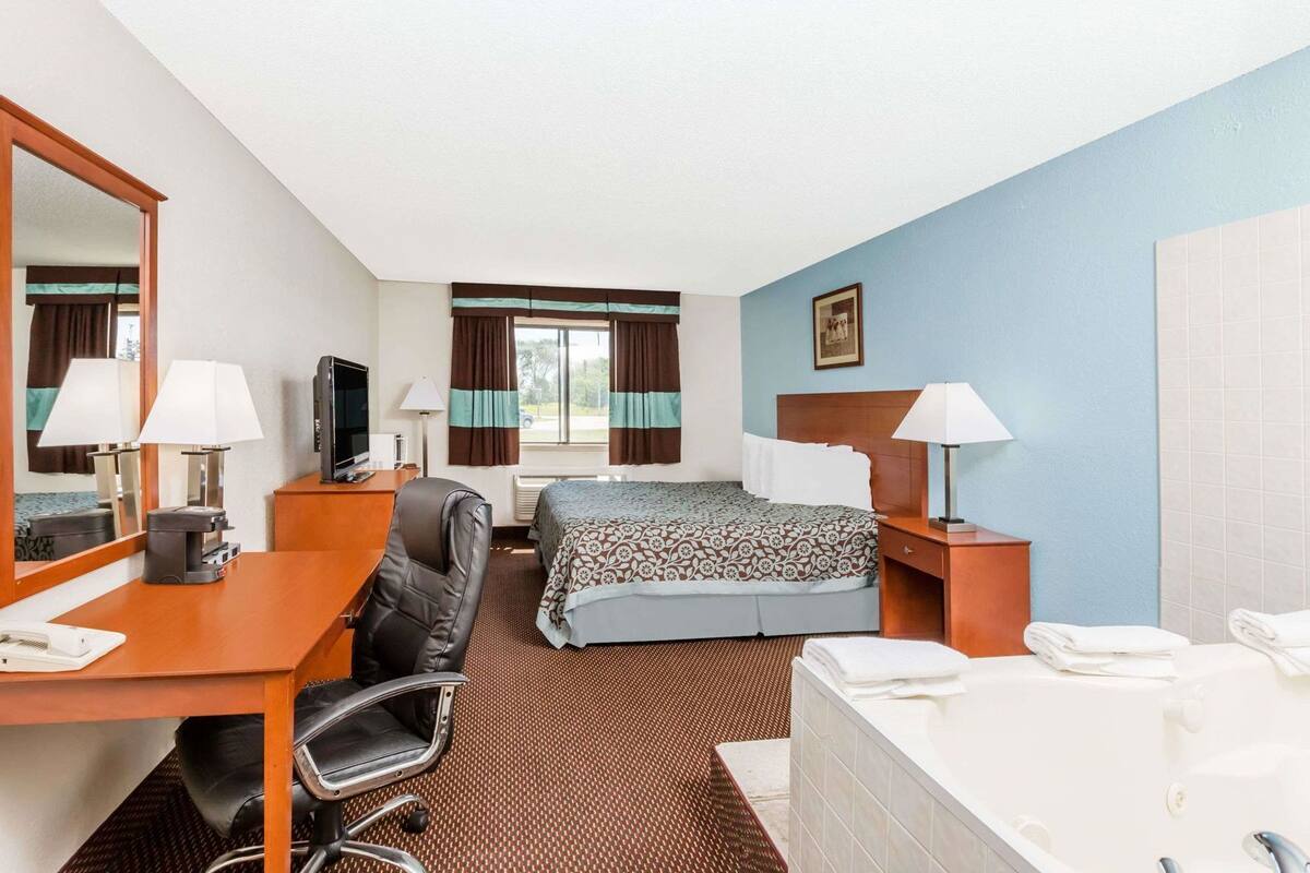 A hotel room with a bed, a desk with a chair, a television, a bathroom with a bathtub, and a window with curtains.
