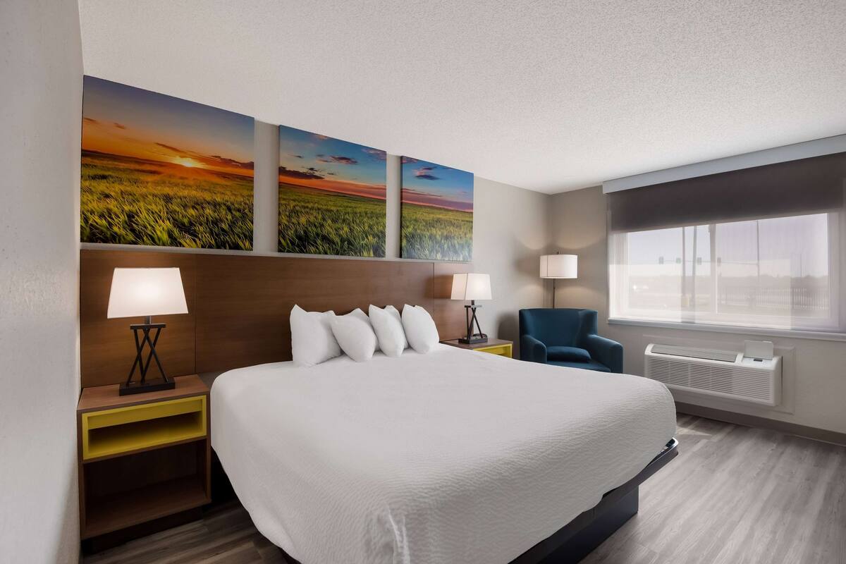 A hotel room with a large bed, two bedside tables with lamps, a chair, and three wall-mounted paintings of a sunset over a field.