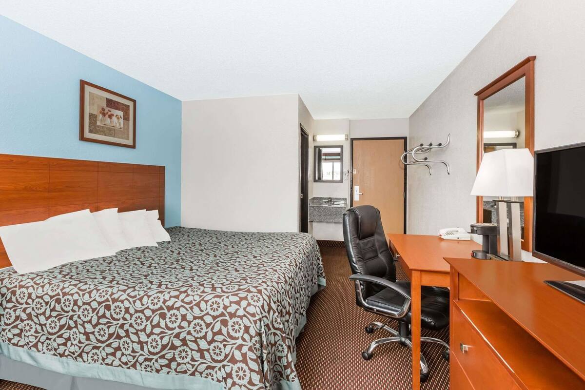 A hotel room with a bed, a desk, a chair, a TV, a lamp, and a bathroom visible through an open door.