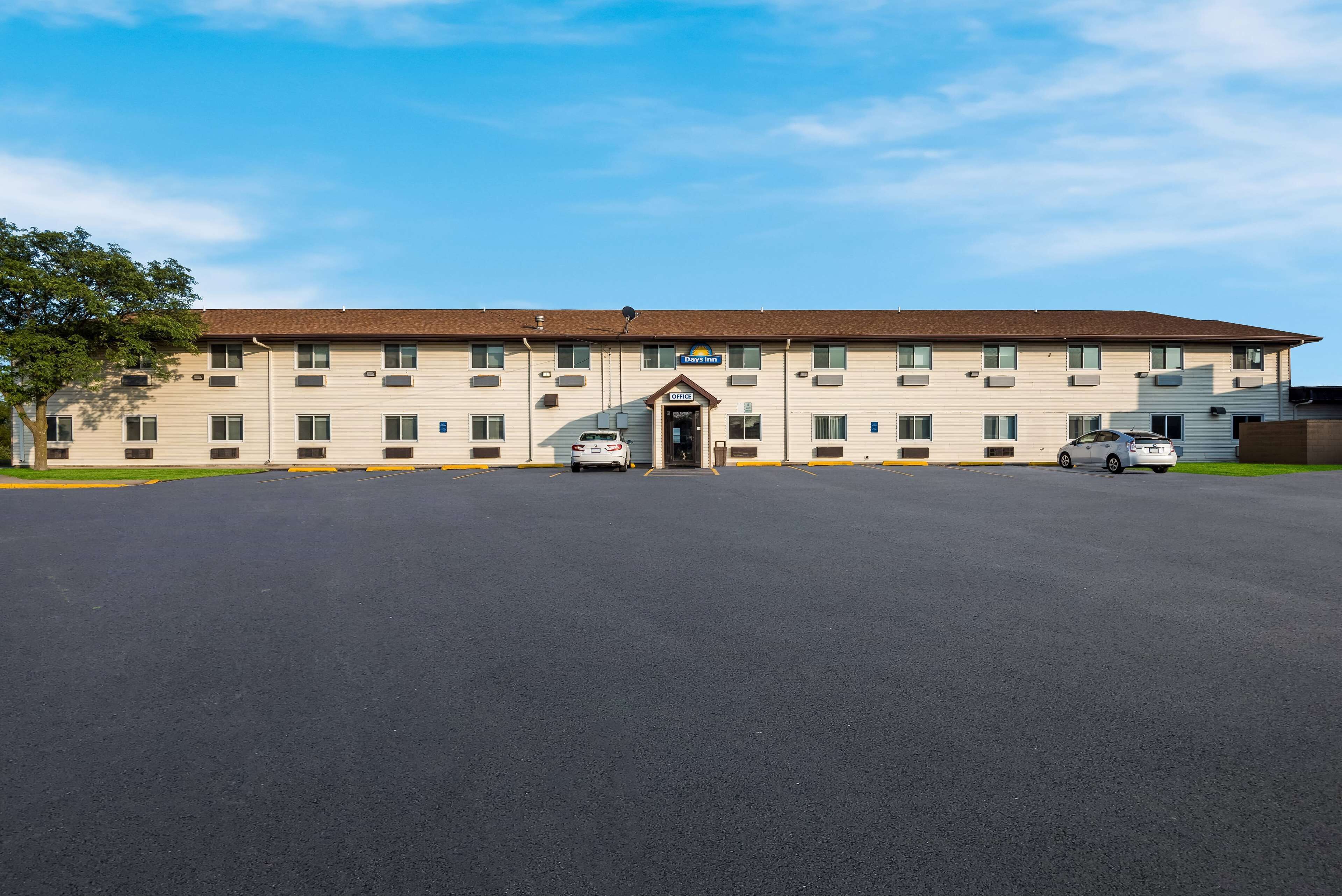 Een meerhoog hotel met een centrale ingang, parkeerplaats en een helderblauwe lucht.