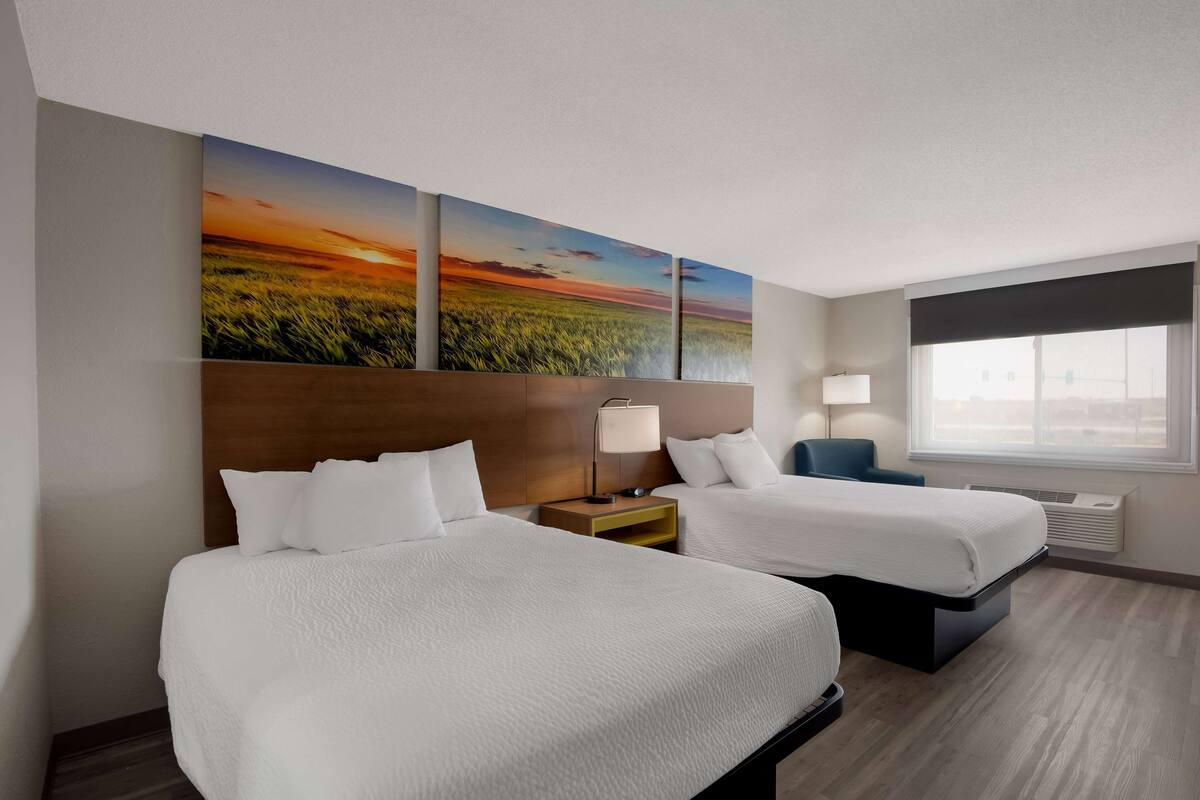 A hotel room with two beds, a nightstand with a lamp, a window with a view, and a wall with three paintings.