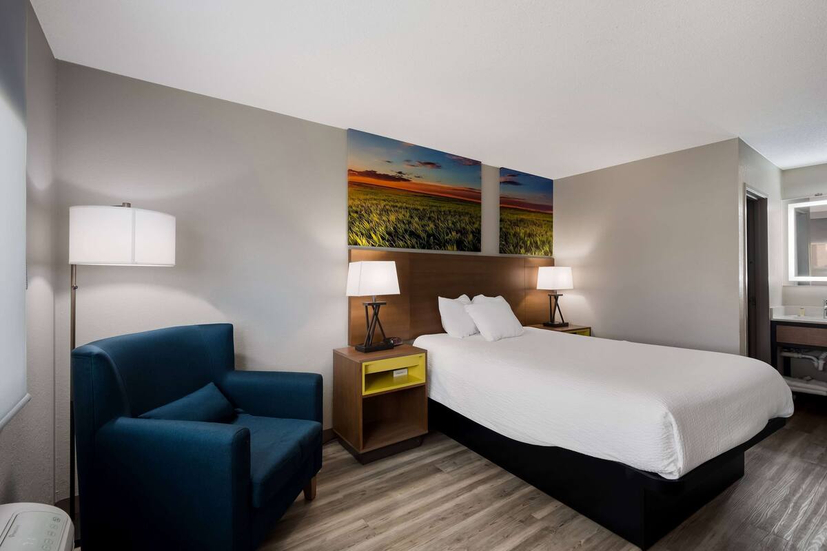 A hotel room with a bed, two bedside lamps, a blue armchair, a wooden nightstand, and two wall-mounted paintings.
