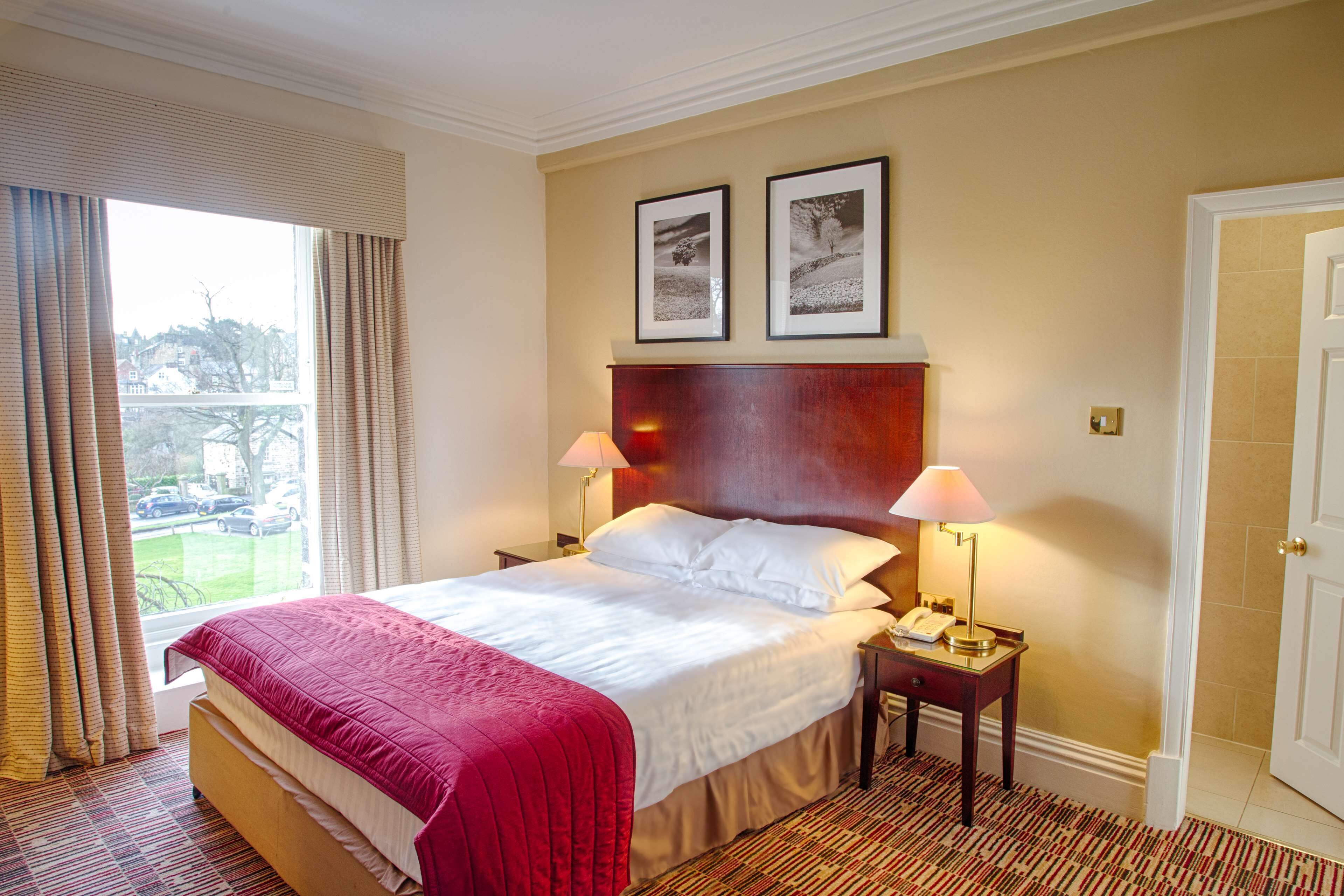 A hotel room with a large bed, two bedside lamps, a headboard with framed pictures, a window with curtains, and a door.