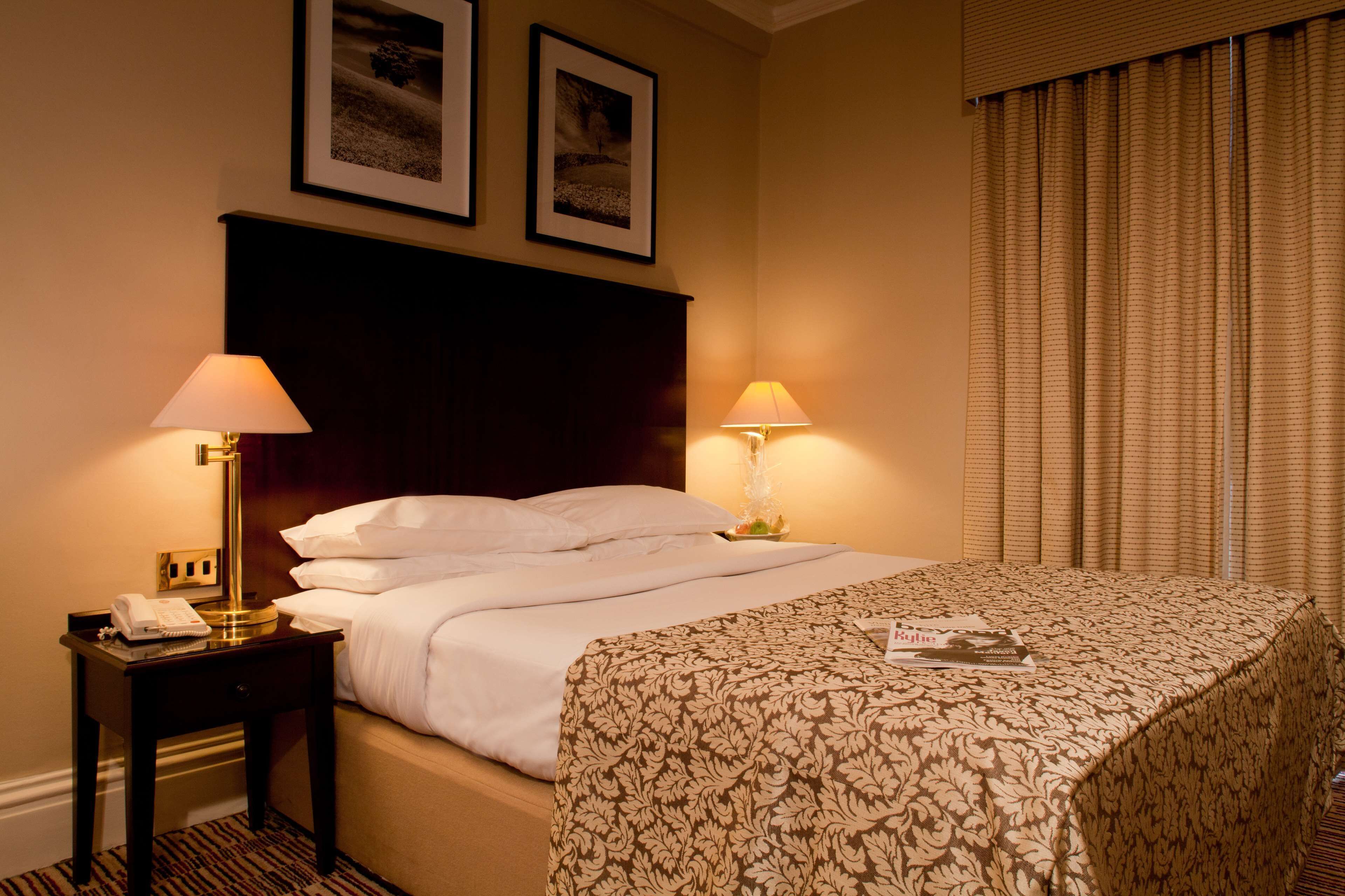 A hotel room with a bed, two bedside lamps, a nightstand with a telephone, and two framed pictures on the wall.