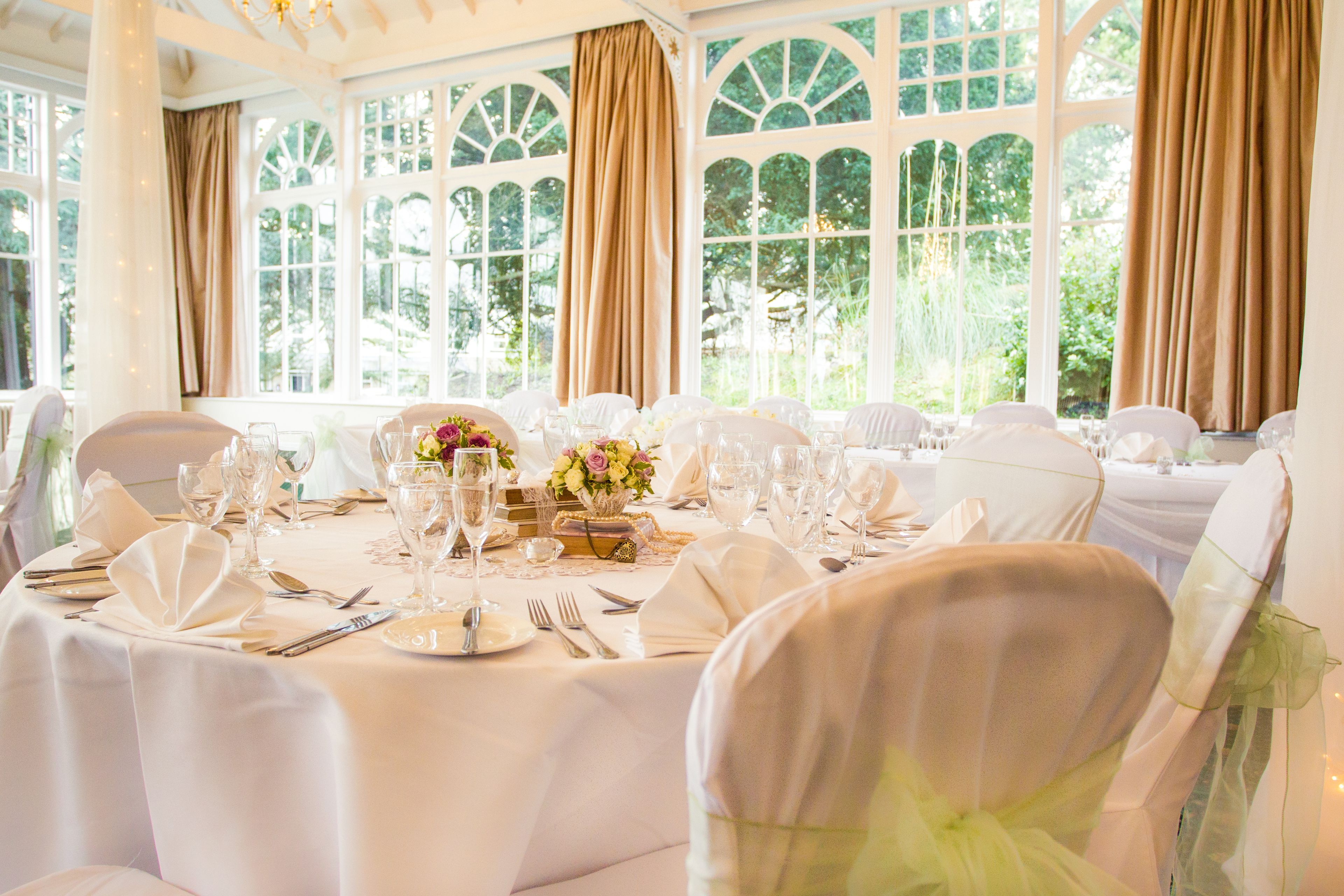 A banquet hall with round tables set for a formal event, large windows with curtains, and a view of greenery outside.