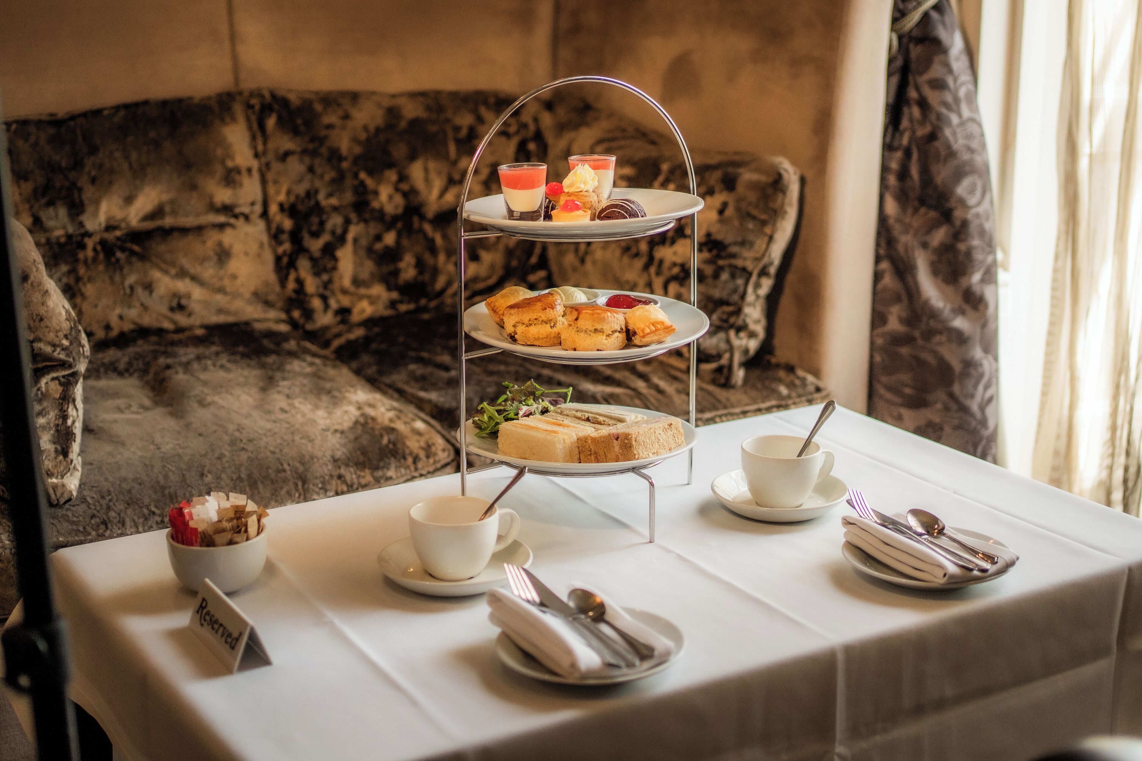 A three-tiered dessert stand with pastries and a cup of tea on a table in a room with a patterned sofa.